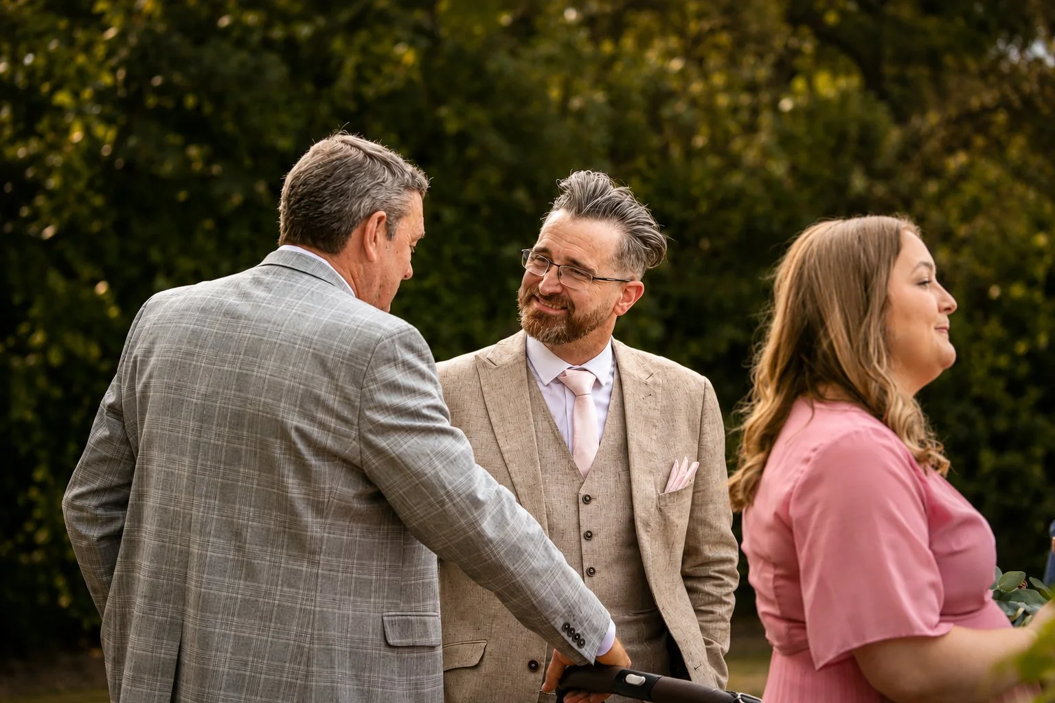 Three people conversing outdoors, two men and one woman, dressed in formal attire, with trees in the background.