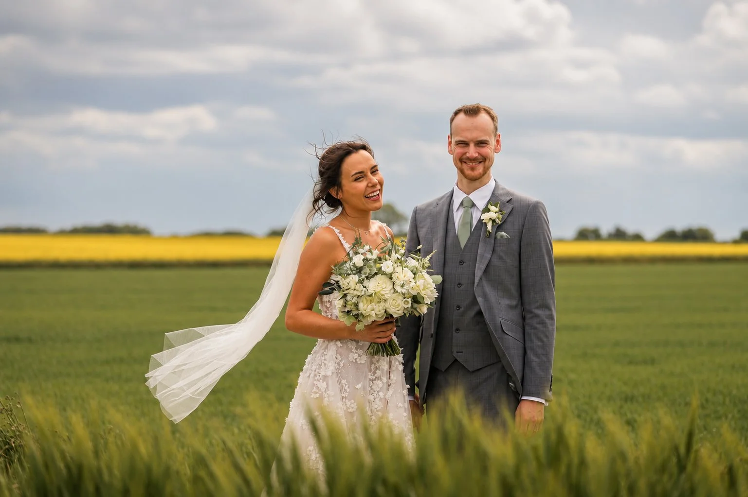 A bride and groom standing outdoors in a green field, smiling, with the bride holding a bouquet of white flowers and wearing a white wedding dress and veil, while the groom is in a gray suit with a tie and boutonniere.