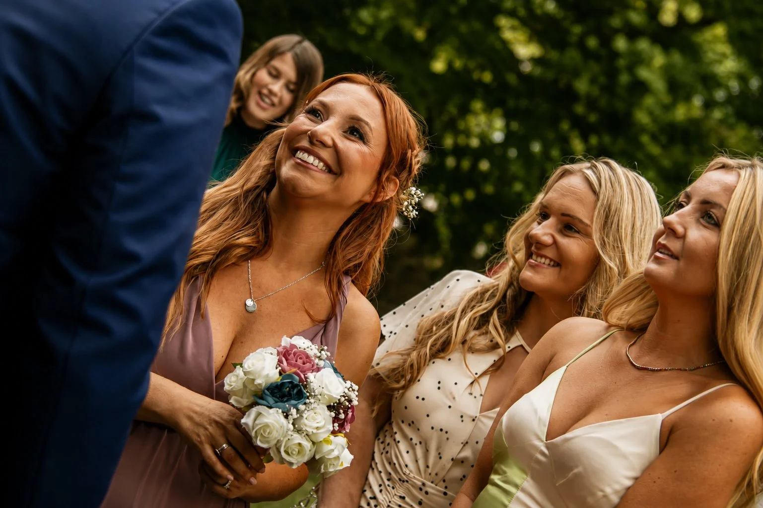 A group of women at an outdoor wedding, smiling and looking at a man in a blue suit, with one holding a bouquet of flowers.
