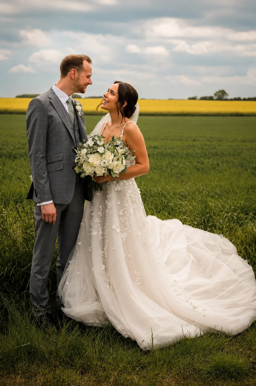 A bride and groom standing in a grassy field, facing each other and smiling. The bride is holding a bouquet of white flowers, and the groom is wearing a gray checkered suit. The bride's wedding dress has lace details and a long train. The background 