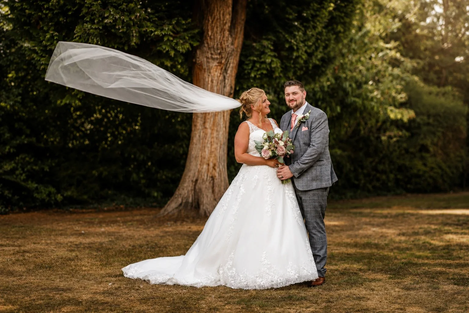 A bride and groom stand outdoors in front of a large tree, smiling and holding a bouquet of flowers. The bride is wearing a white wedding dress with lace details, and the groom is dressed in a gray suit with a pink tie and boutonniere. A long veil is