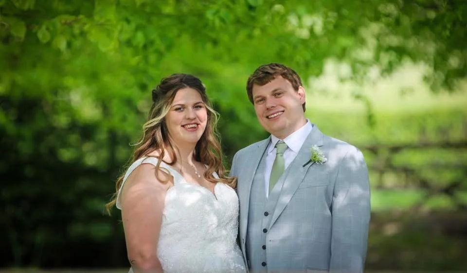 A bride and groom standing outdoors in a lush green park, smiling at the camera during their wedding.