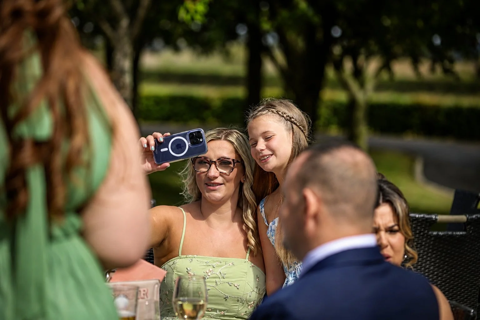 A group of people gathered outdoors, taking a photo together, with a woman in a green dress and a girl with braided hair smiling, and others around them.