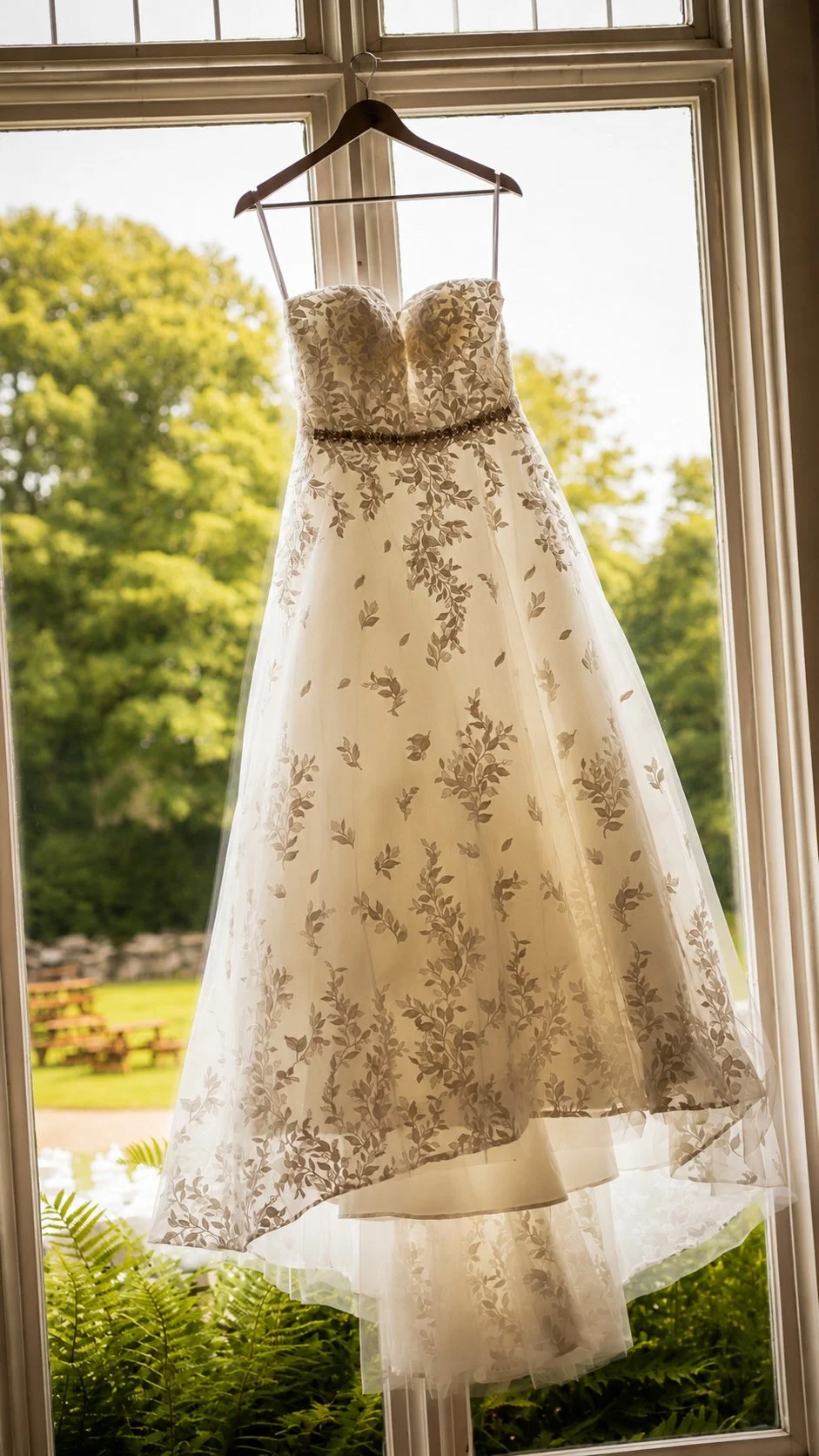 A wedding dress with a floral pattern hanging on a hanger in front of a window with a view of green trees and a garden outside.