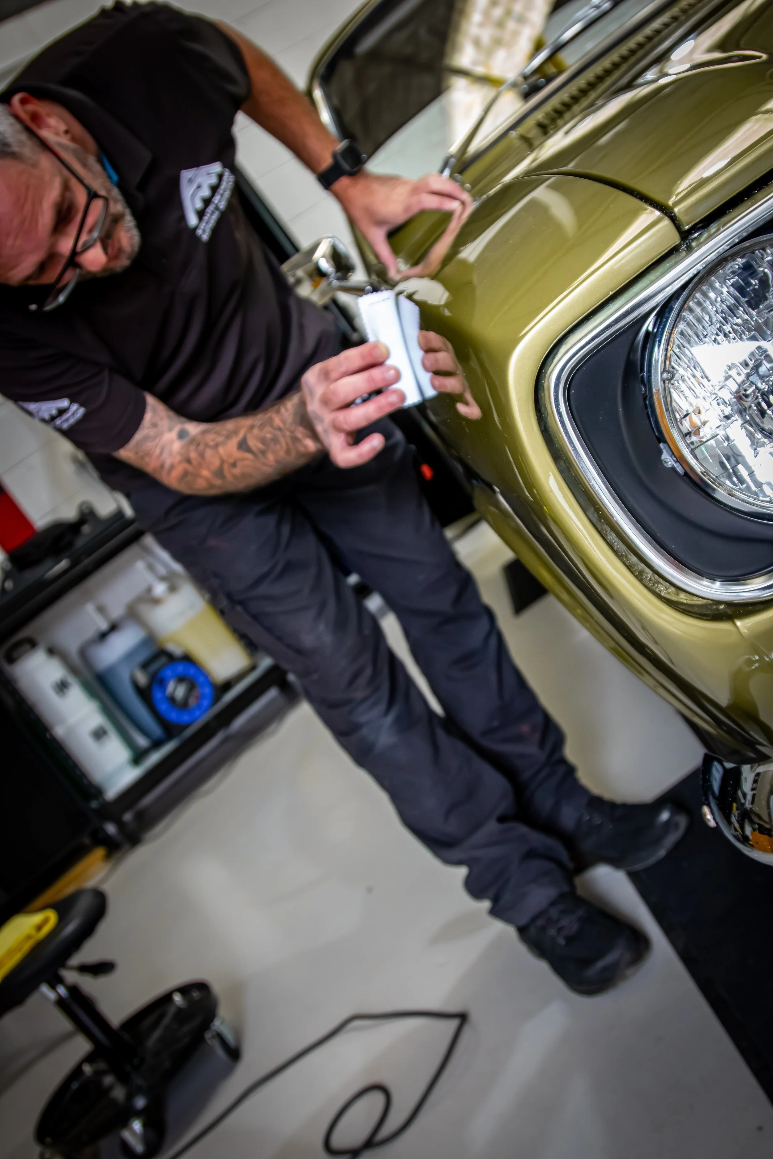 A person with tattoos on their arm, glasses, and a black shirt is working on a green car, possibly attaching or inspecting the front hood. The person is in a garage or workshop with tools and equipment visible in the background.