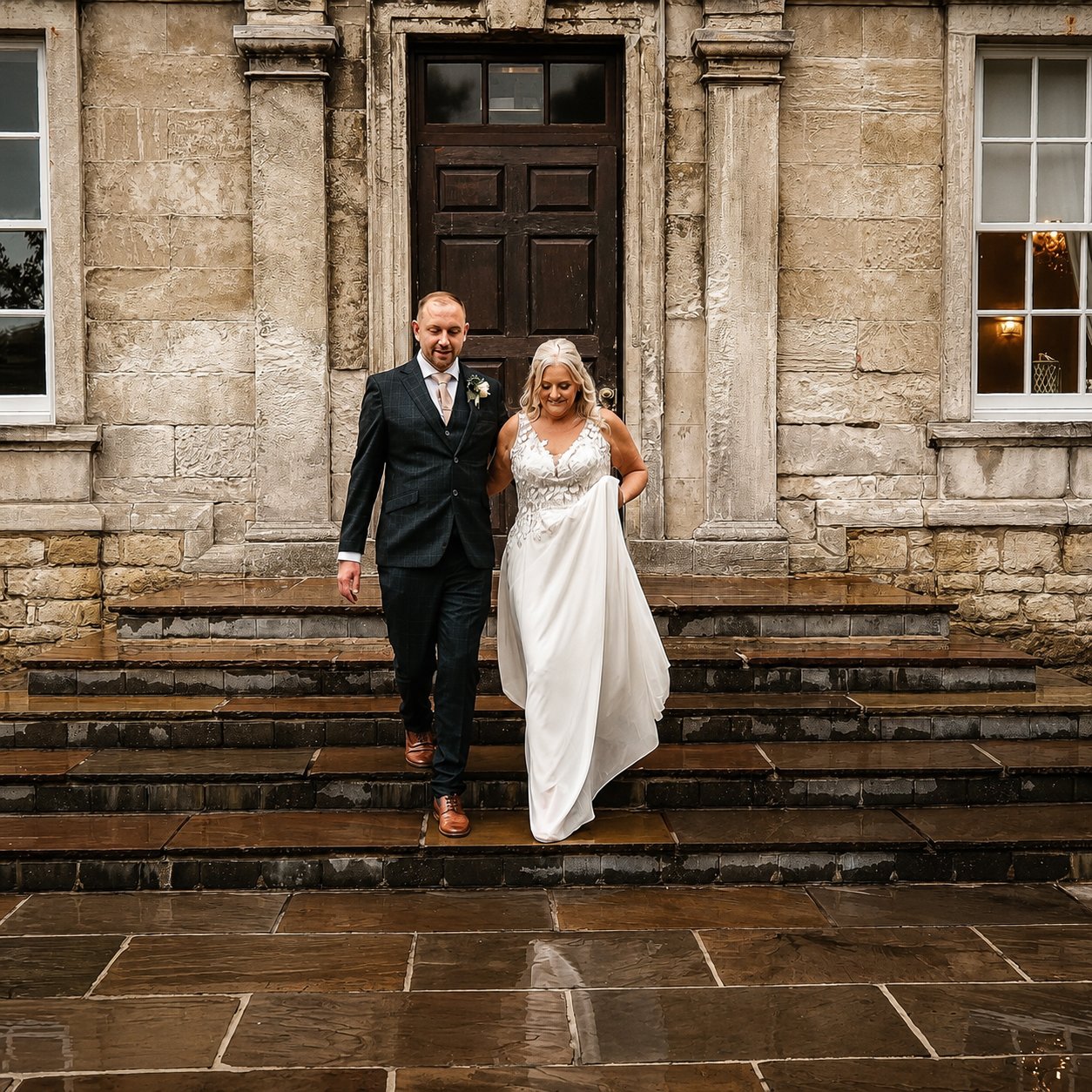 A bride and groom walking down stone steps outside a historic building with large windows and a wooden door.