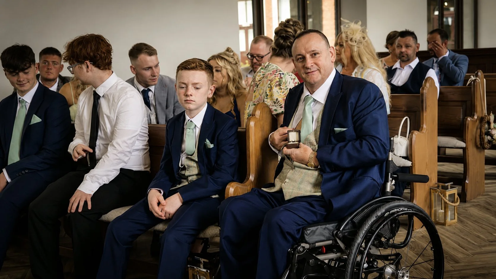 A group of people seated in a church during a wedding. In the front row, a man in a wheelchair holds a ring box, next to a young boy. Others around them are dressed in formal attire, some looking forward and others engaged in conversation.