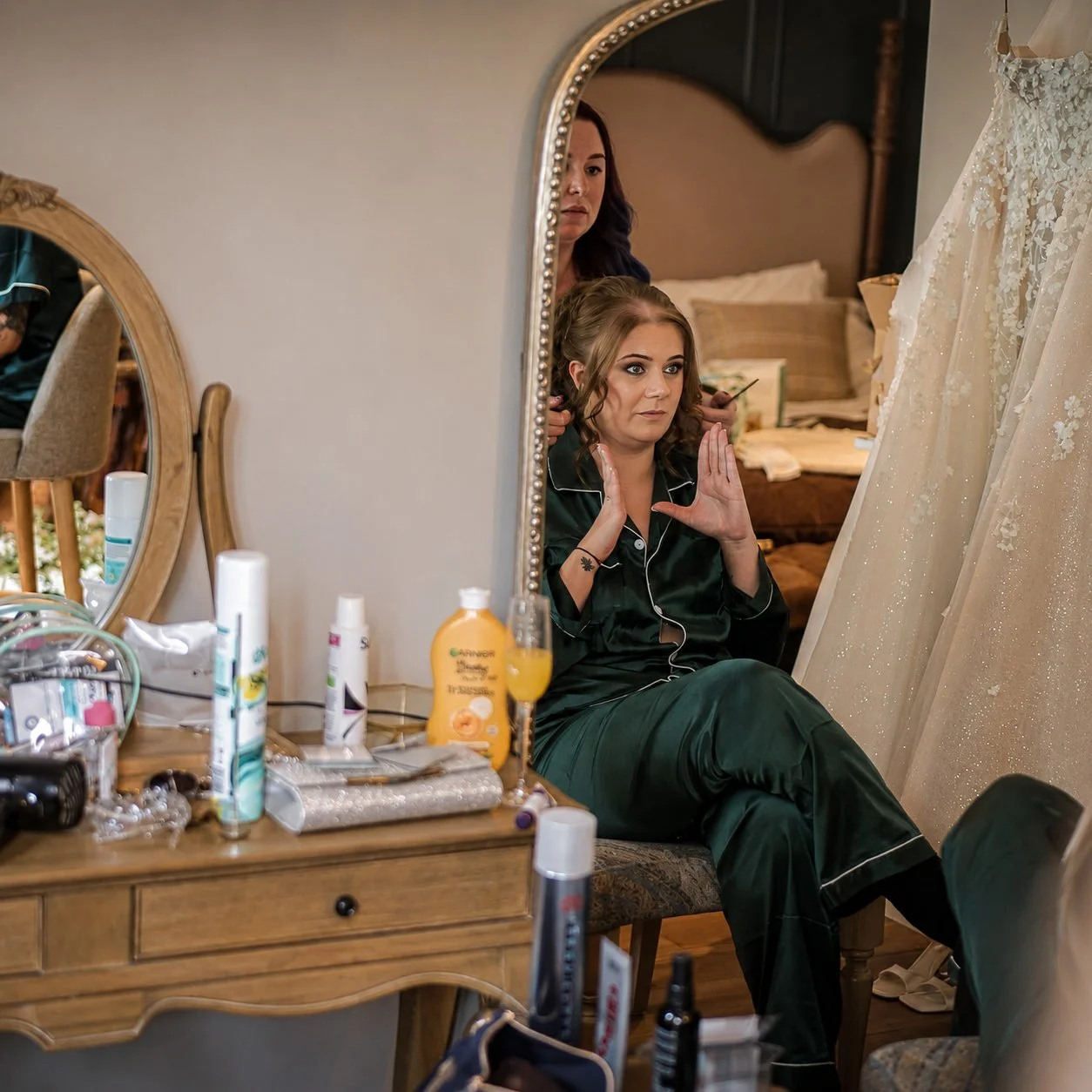 A woman sits in front of a mirror adjusting her hair, getting ready, while dressed in dark green satin pajamas. The mirror reflects her face and part of her bedroom. The dresser in front of her holds various toiletries, makeup, and personal items, in