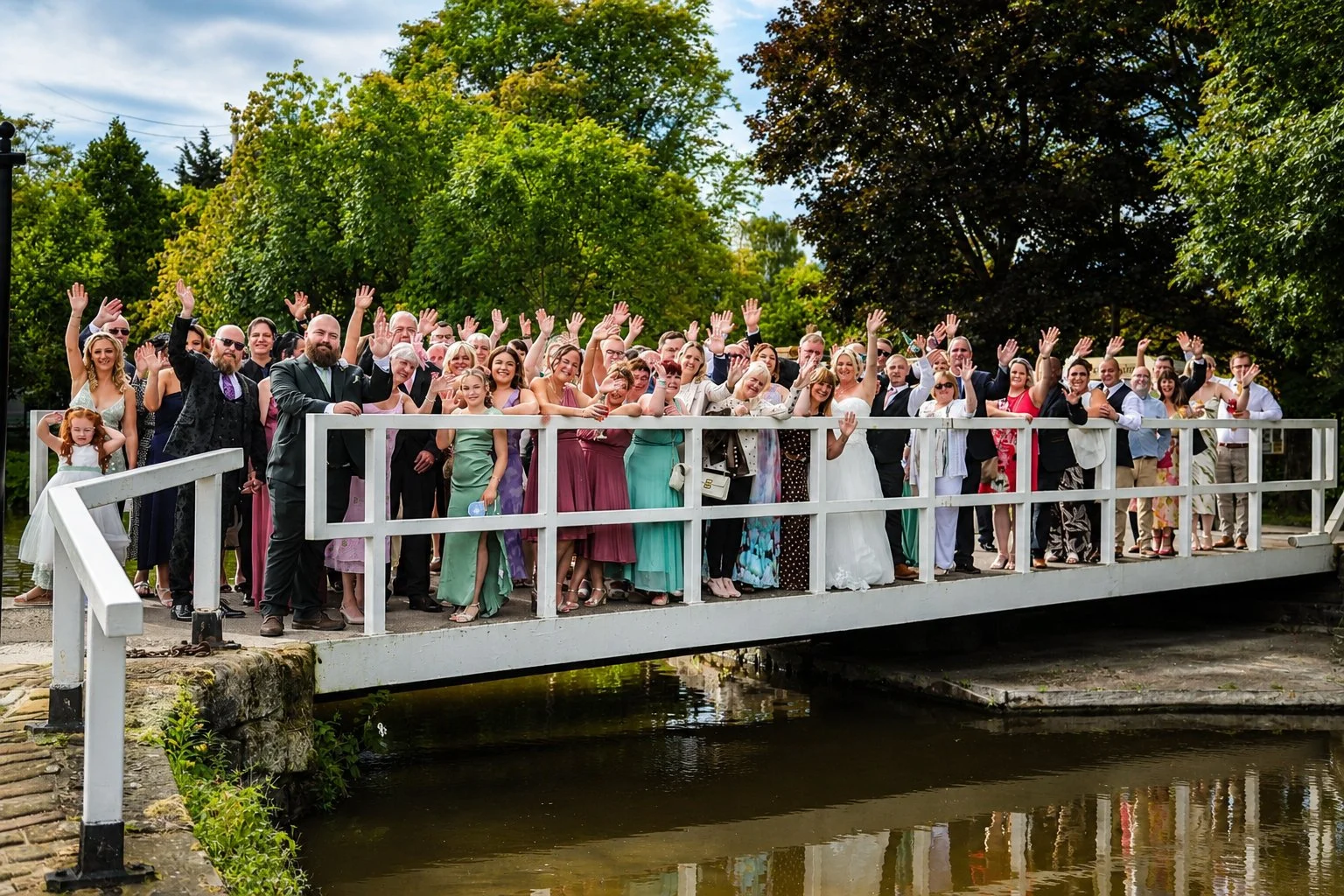 A large group of people on a white bridge over water, waving and smiling, with trees in the background, dressed in formal and semi-formal attire, at a celebratory event.