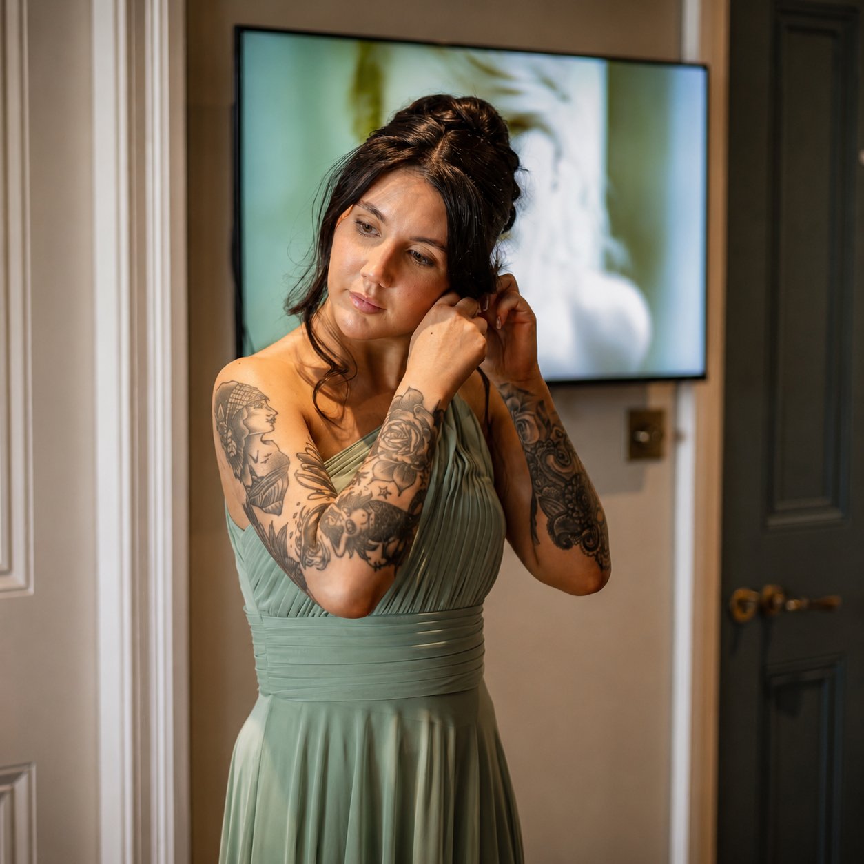 A woman with black hair and tattoos is adjusting her earring while wearing a green dress in a room with beige walls and a television in the background.