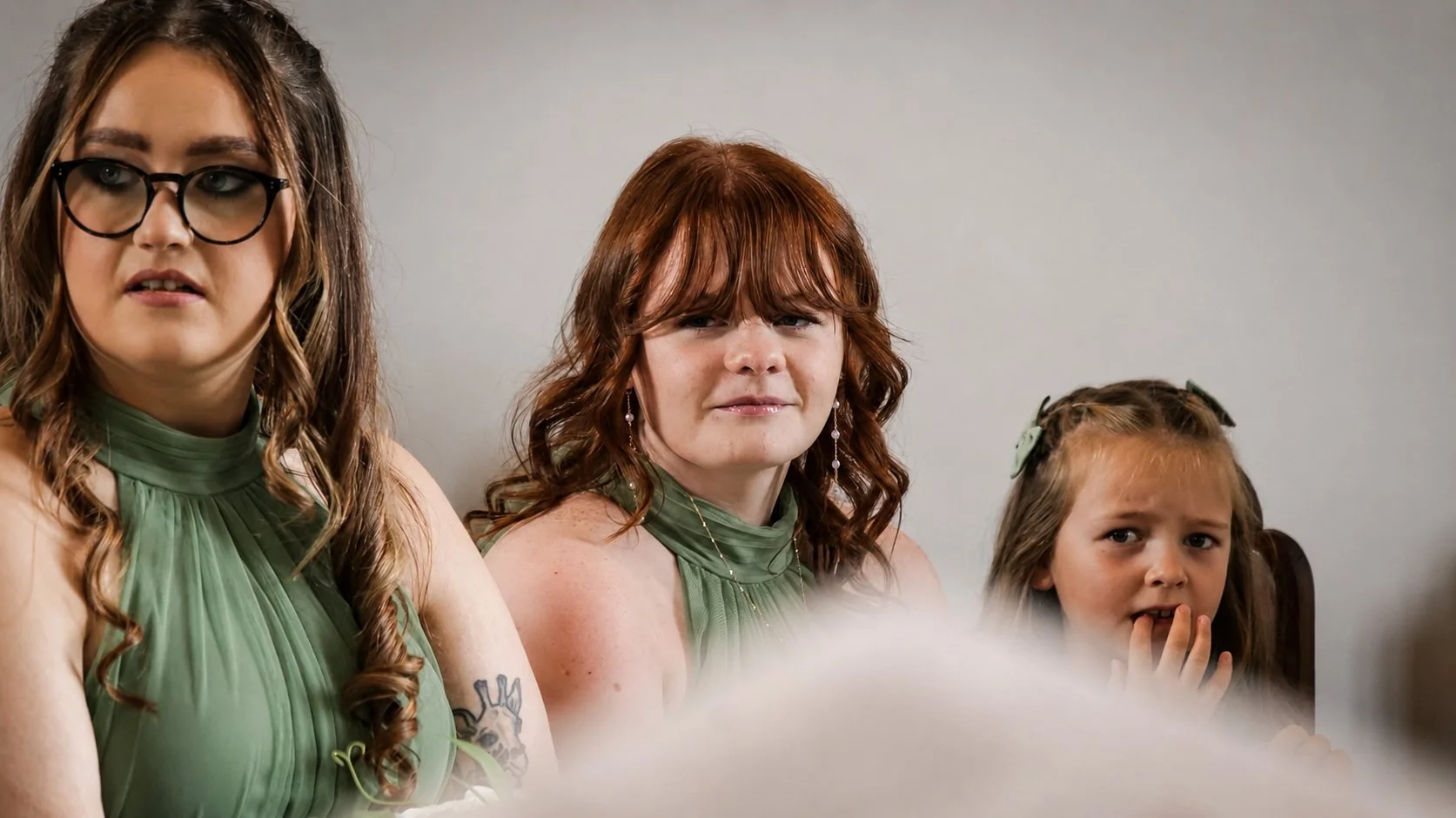 Three women and a young girl sitting together, the women wearing green dresses, with varied expressions, against a plain background.
