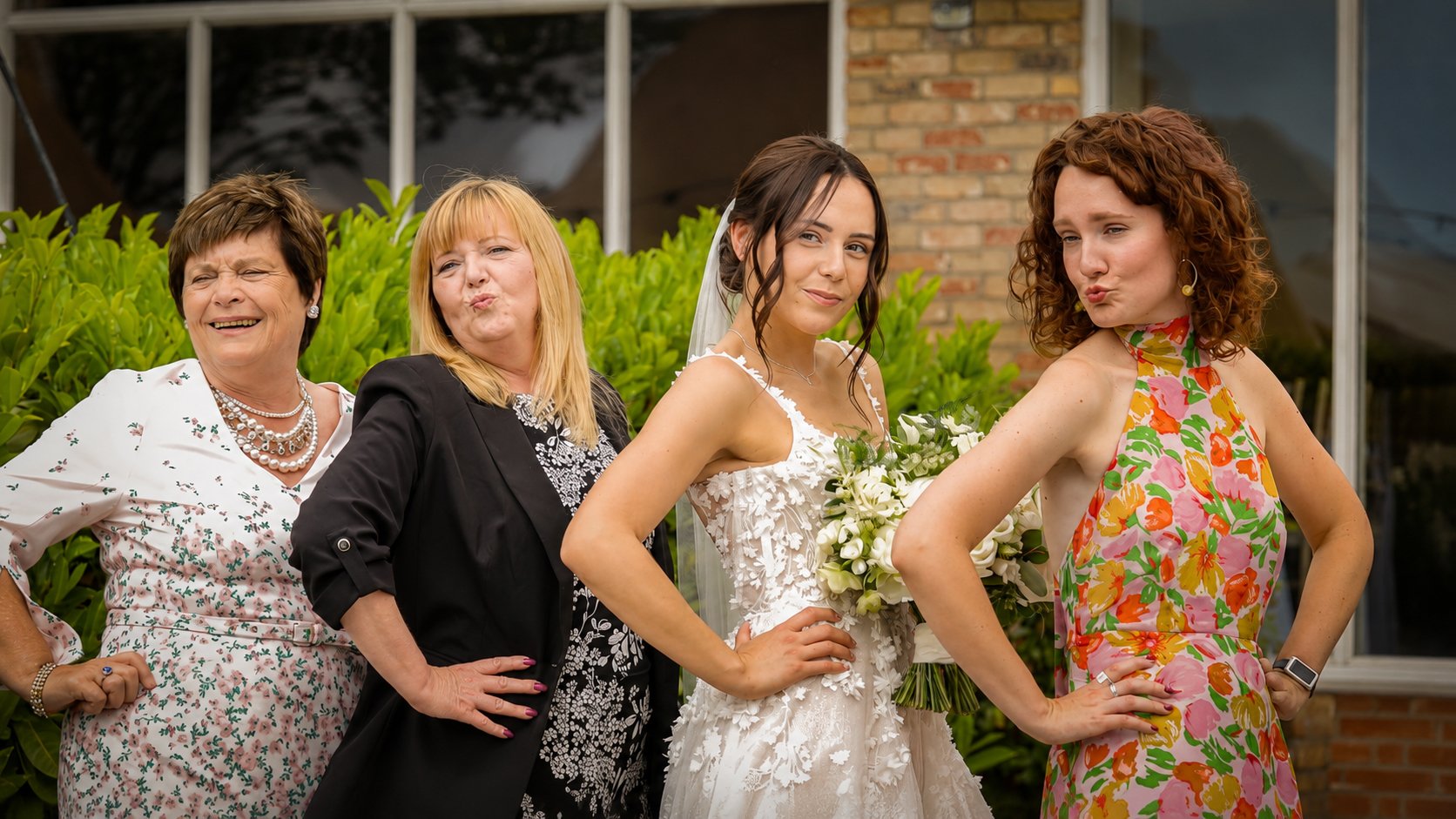 Four women standing outdoors, with a bride in a white wedding dress holding a bouquet, posing with two women in colorful outfits, all making playful faces, in front of green plants and a brick building.