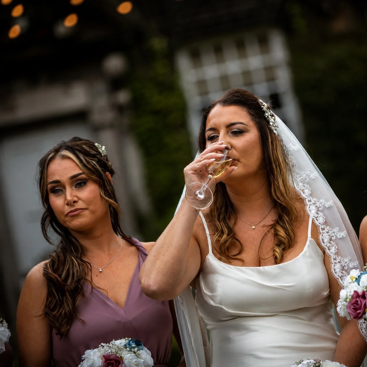 A bride in a white wedding dress with lace details on her veil is drinking from a glass of champagne, standing next to a bridesmaid in a lavender dress at an outdoor wedding reception.