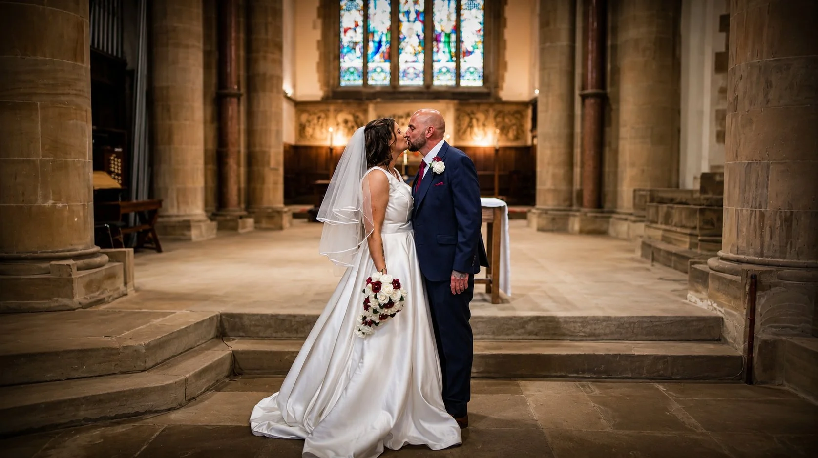 A bride and groom kiss inside a church, with stained glass windows behind them and stone columns on either side.