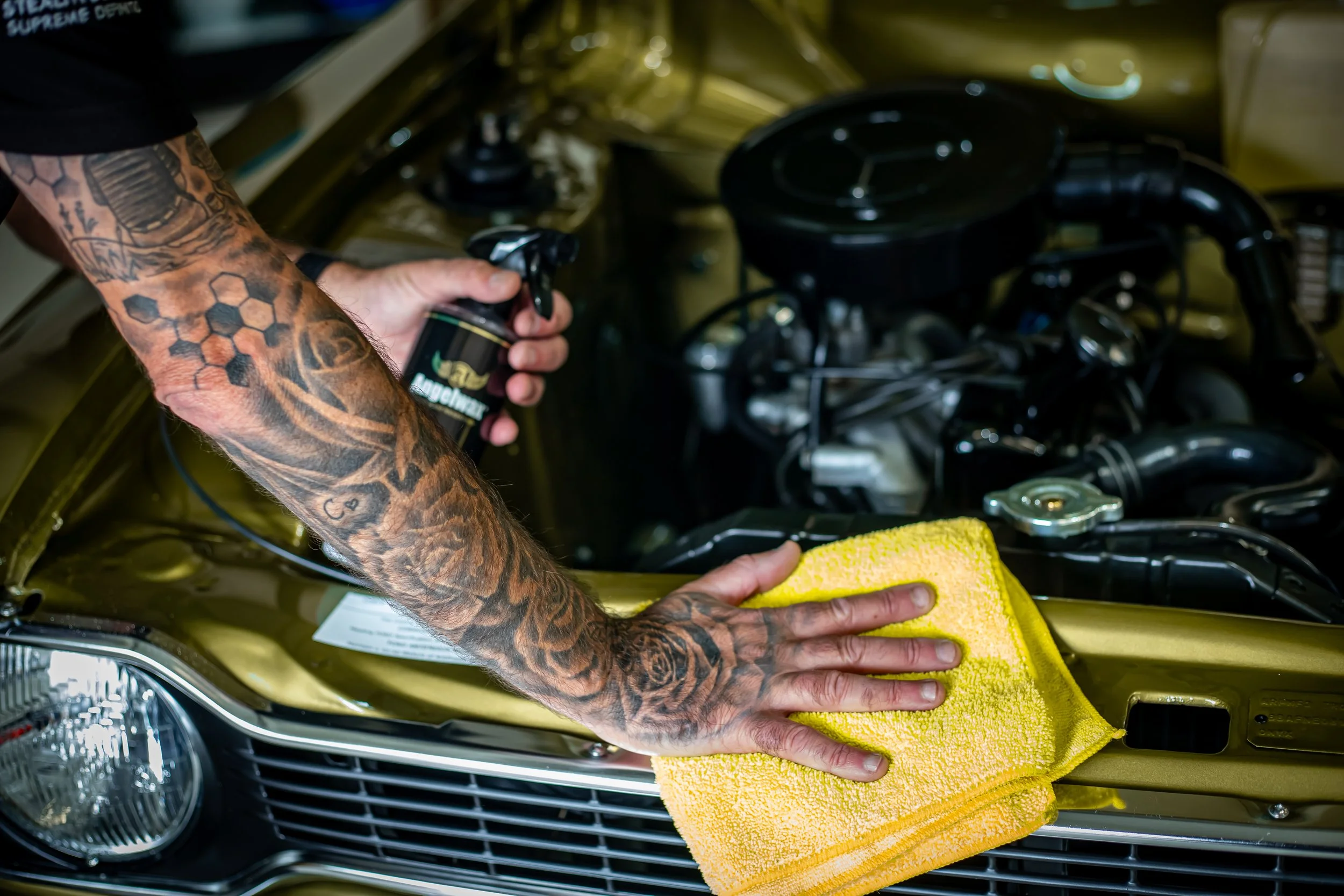 Person with tattooed arms cleaning the front of a gold-colored car with a yellow microfiber cloth.