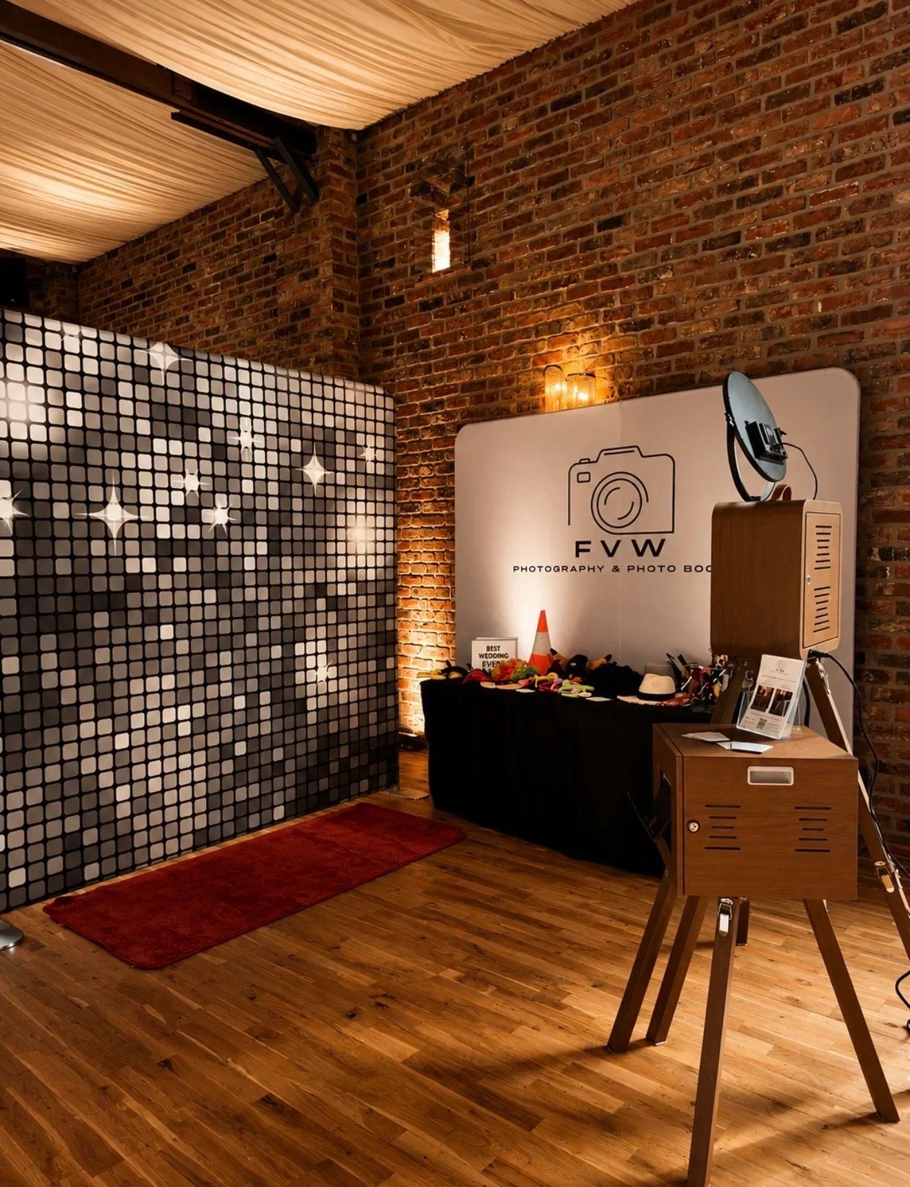 Indoor photo of a photo booth setup with a brick wall background, a black table with props, and a sign that reads 'FVW Photography & Photo Booth.' A camera prop stands on a wooden tripod in the foreground, and a decorative partition with star patterns is on the left side. Warm lighting highlights the scene.