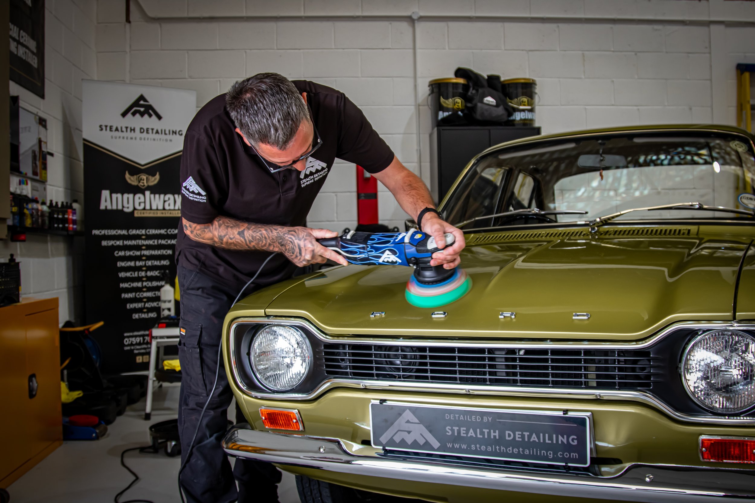 A man polishing a vintage green car with a polishing machine in a garage, with a banner for Stealth Detailing in the background.