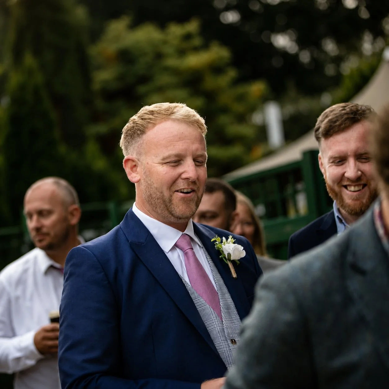A man in a blue suit with a pink tie and a white flower boutonniere smiling at a wedding ceremony outdoors.