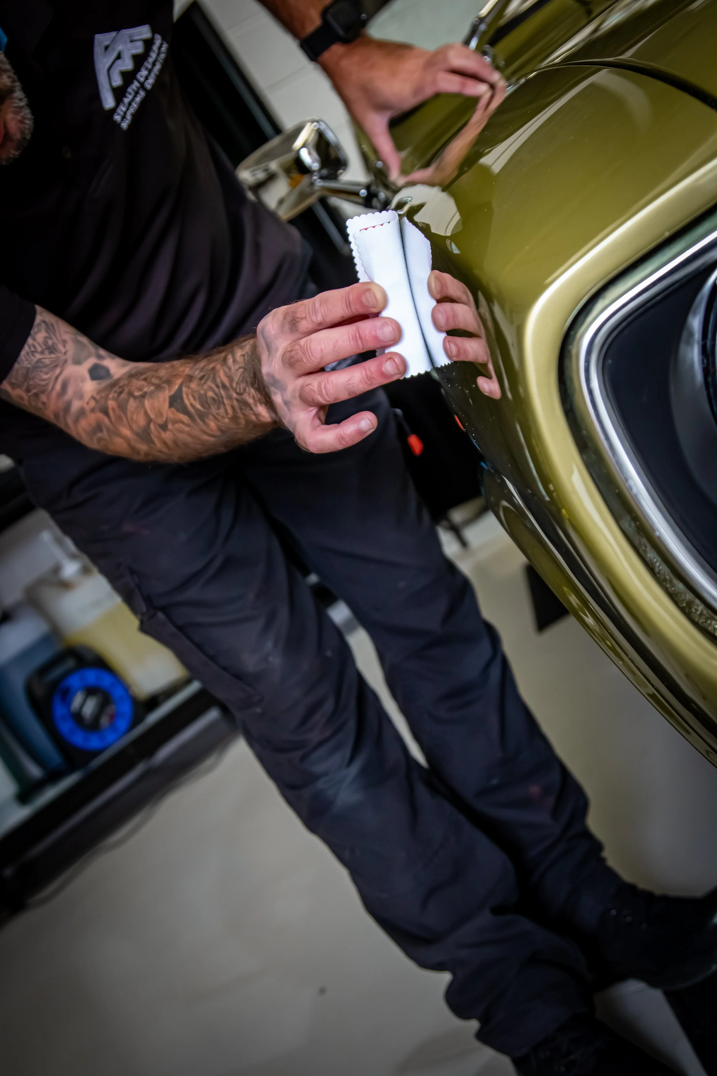 Person polishing a green car with a white cloth, in a garage setting with tools and equipment visible in the background.