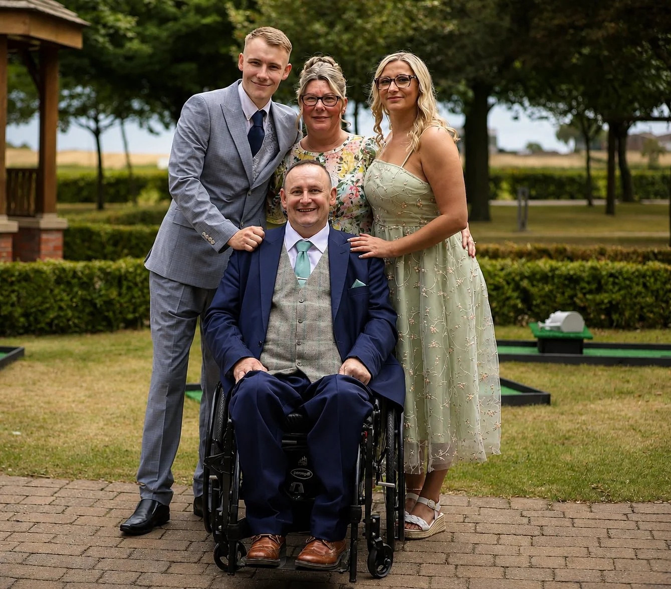 A group of five people posing outdoors: an elderly man in a wheelchair, a young man in a gray suit, an older woman in a floral dress, and a young woman in a light green dress. They are standing on a brick path with trees and a park in the background.