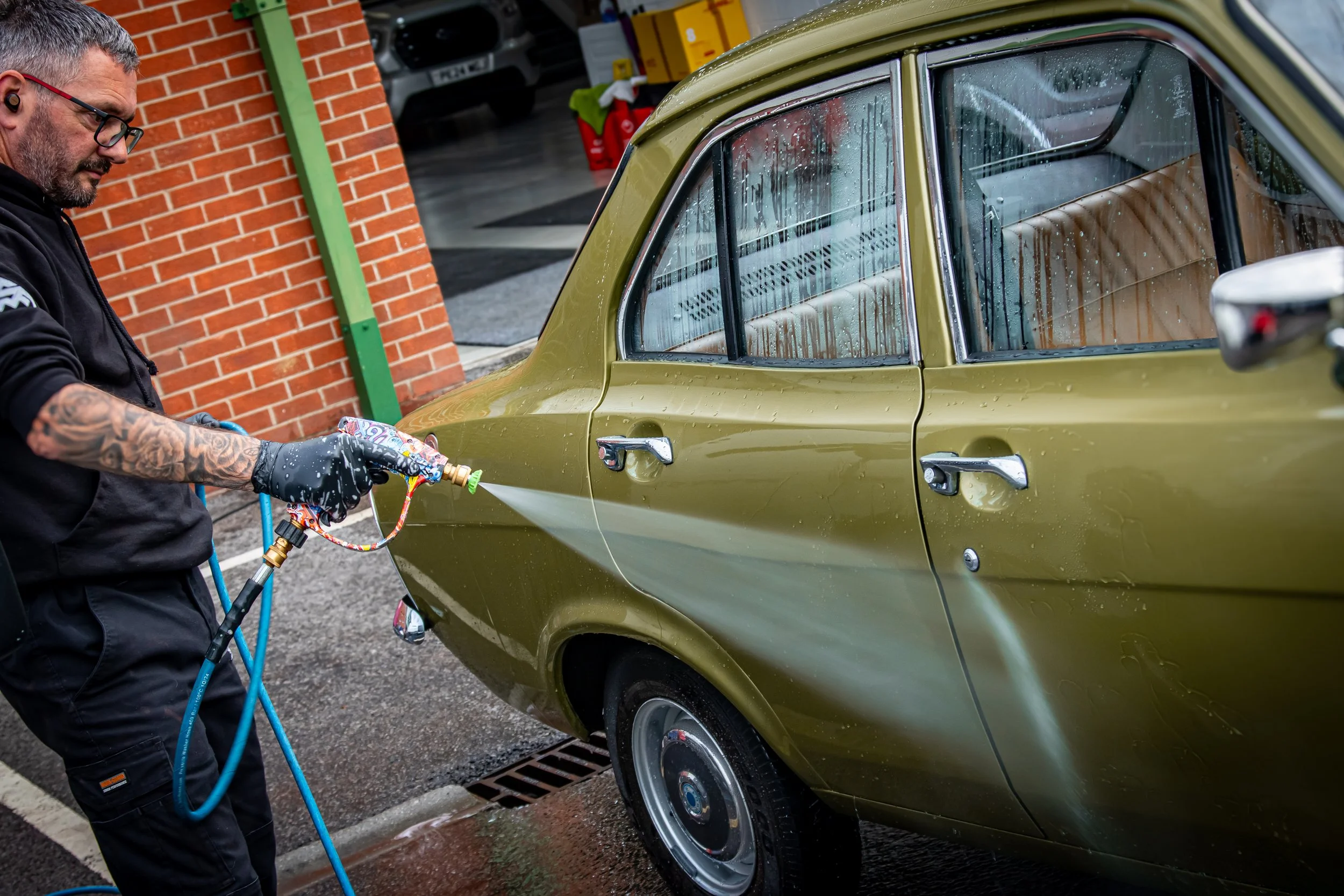Man washing a green vintage car with a hose at a car wash station, rain on the windows, brick wall and parked cars in the background.