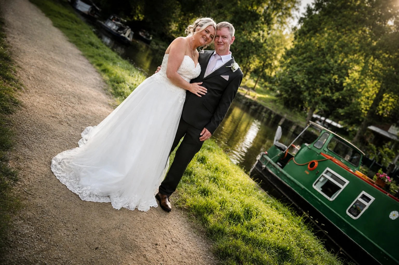 Bride and groom in wedding attire standing on a dirt path beside a canal with a green boat, surrounded by trees in the evening sunlight.