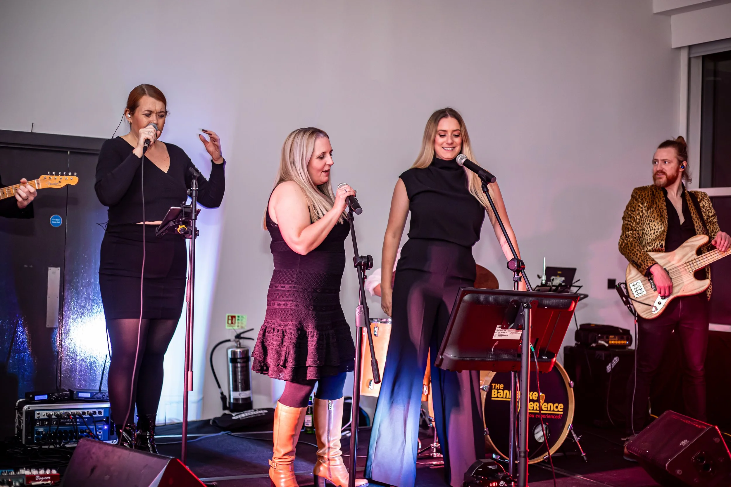 A musical performance featuring four women singing and a man playing bass guitar on stage, with various musical equipment visible.