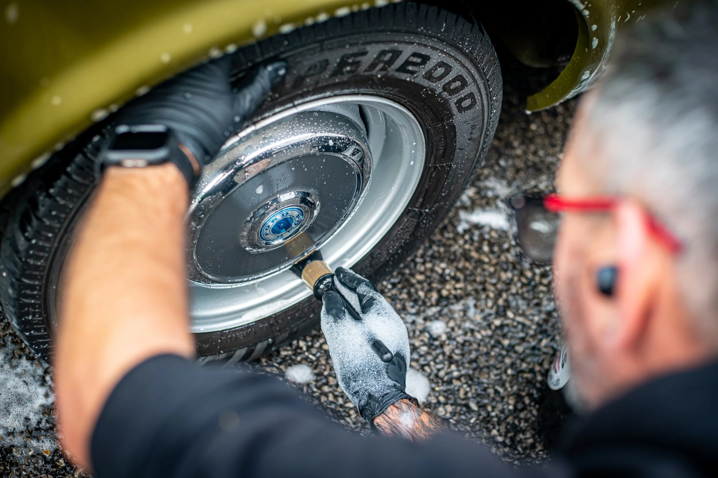 A person washing a yellow car's wheel with water and a sponge while wearing black gloves.