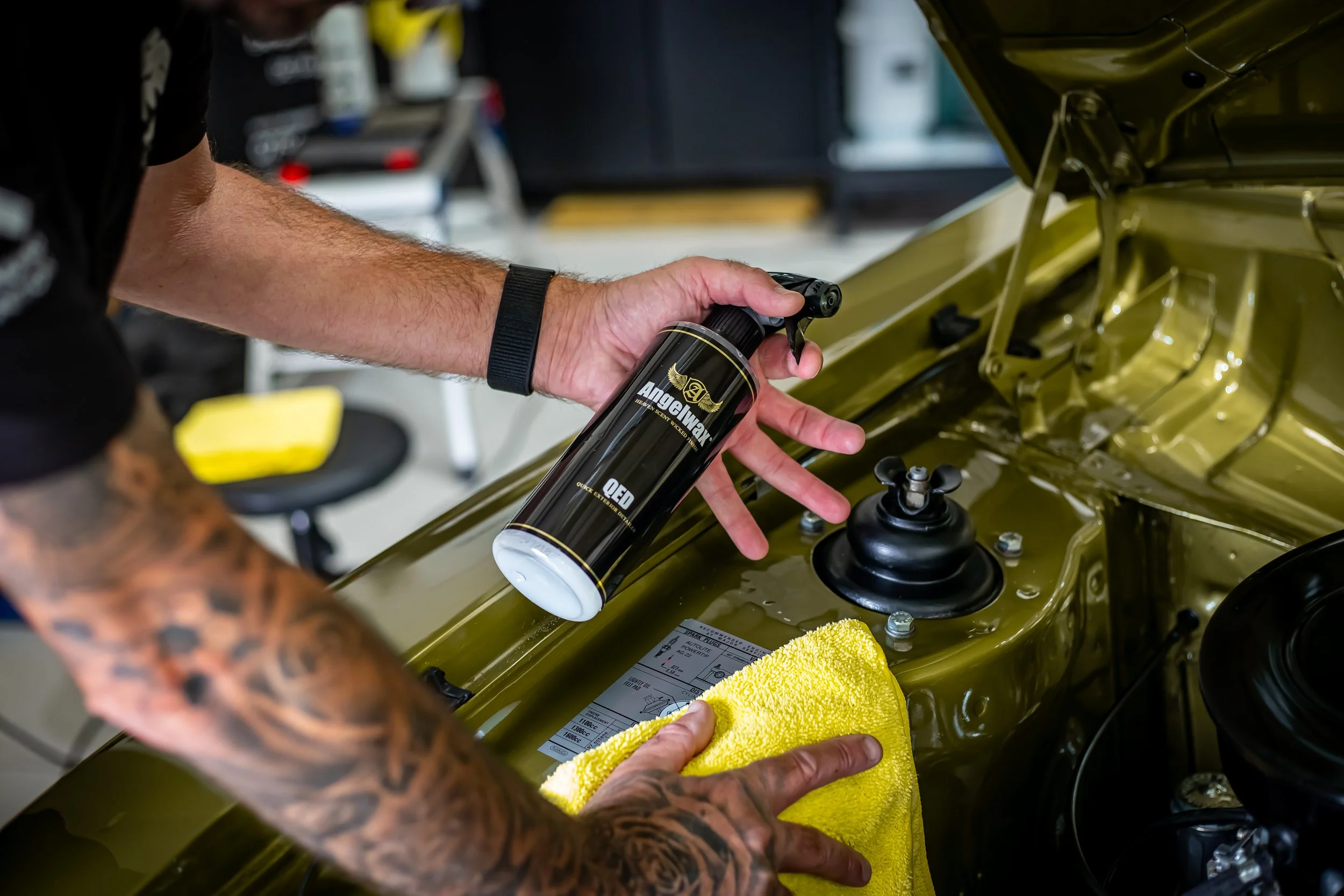 Person spraying a car part with a can labeled 'Angelwax' as they clean or apply something to the vehicle.