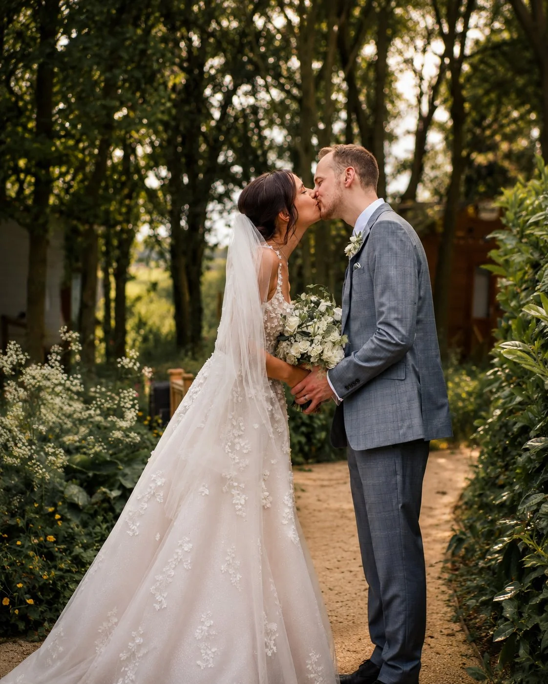 A bride and groom share a kiss outdoors during their wedding, with the bride holding a bouquet of white flowers and wearing a lace wedding gown, and the groom in a gray suit with a floral boutonniere, surrounded by greenery.