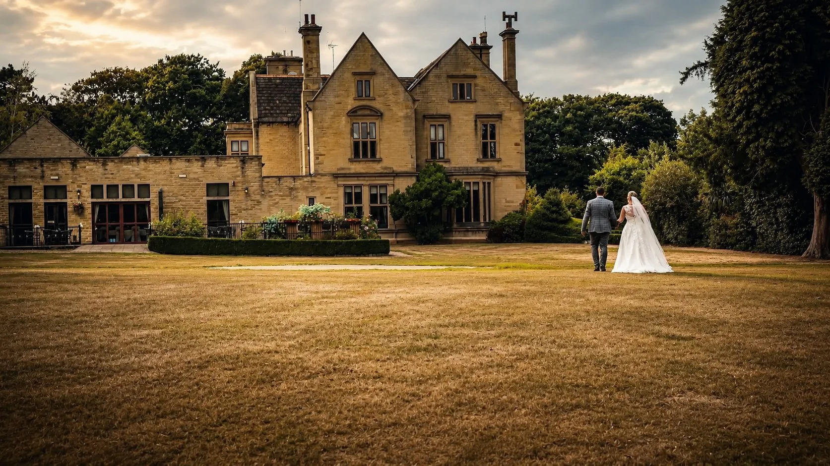 A bride and groom walking hand in hand on a lawn towards a large, historic stone mansion surrounded by trees and greenery with a dramatic sky overhead.
