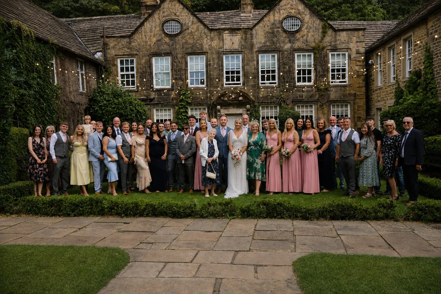 A large group of people gathered outside on a lawn in front of an old stone building with string lights, posing for a group photo at a wedding.