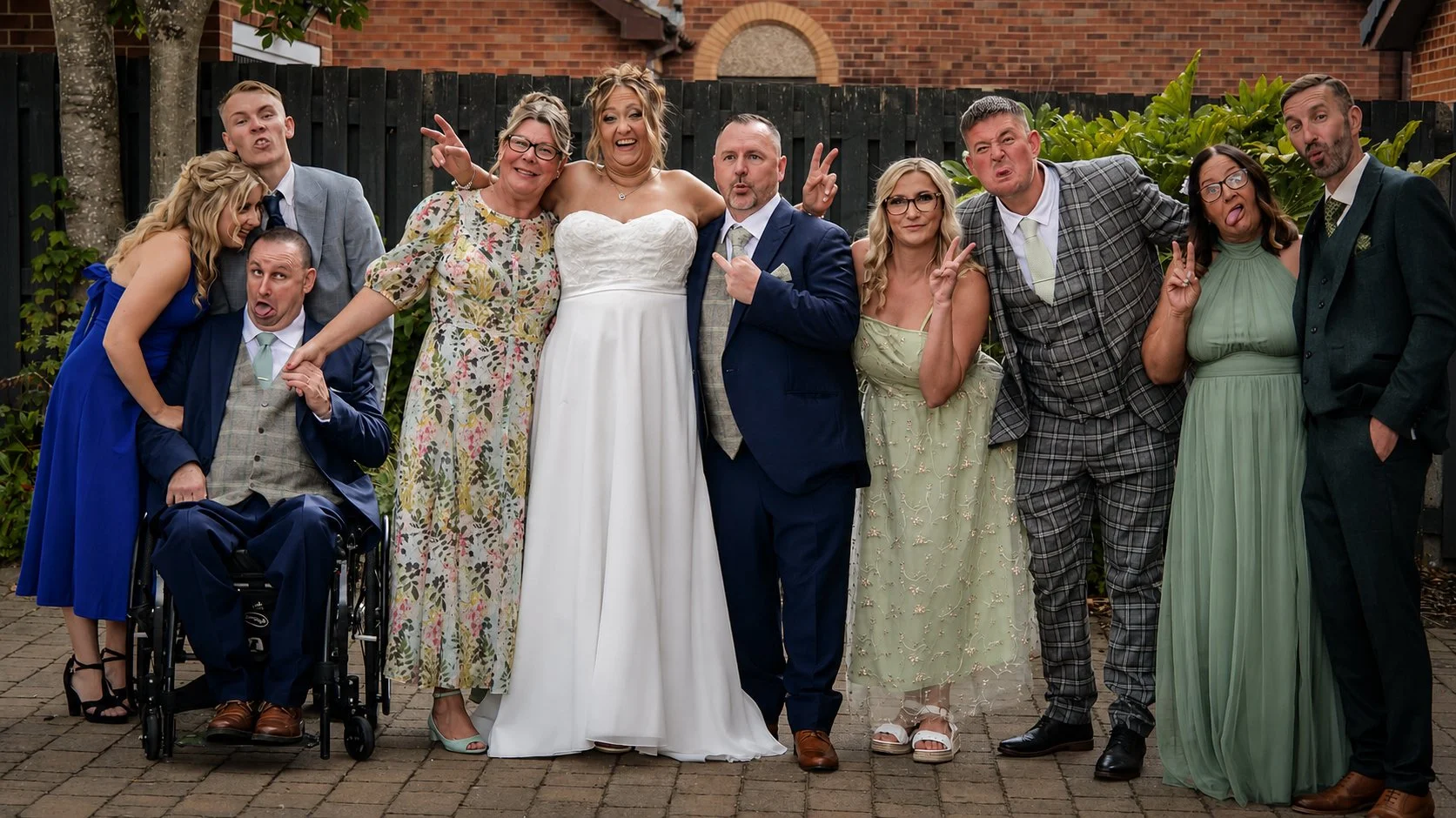 Group of people posing for a photo outdoors, some making funny faces and gestures, including the bride in a white strapless wedding dress, in front of a brick building and a wooden fence.