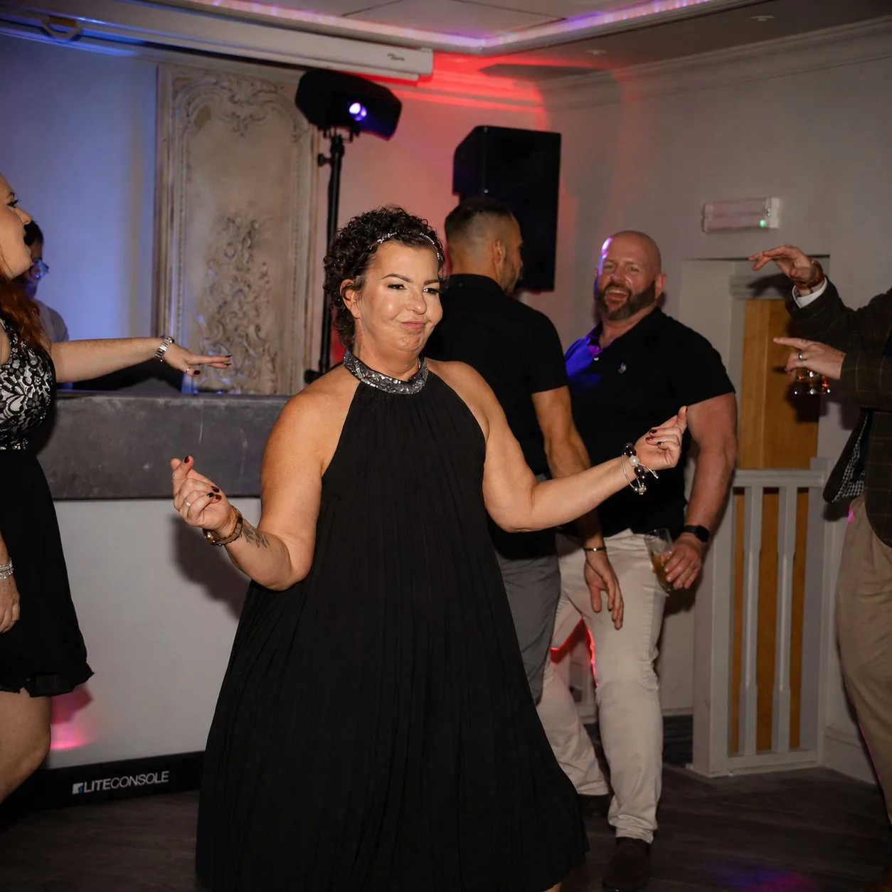 People dancing and having fun at a party or celebration, with one woman in the foreground wearing a black dress, smiling, and swinging her arms to music.