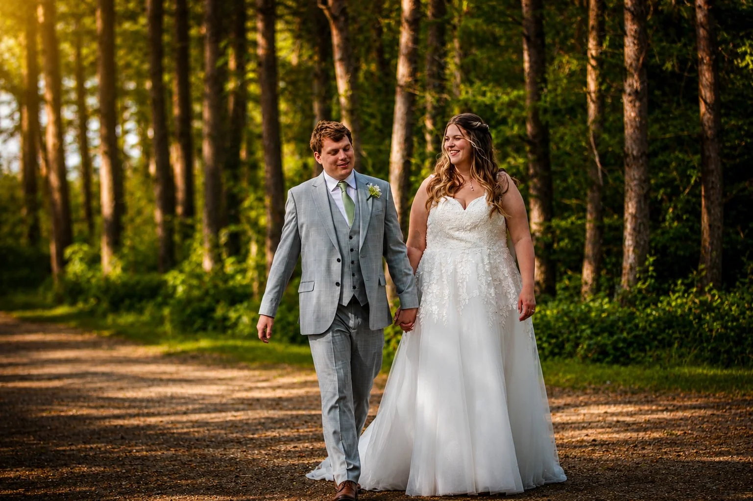 A newlywed couple walking hand in hand through a forested area during sunset, with the bride in a white wedding gown and the groom in a light gray suit.