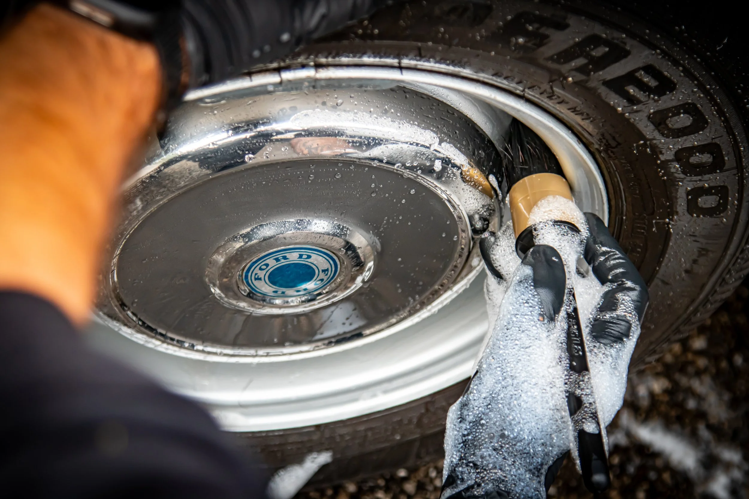 Person washing a car wheel with a hose, soapy water and soap bubbles visible.