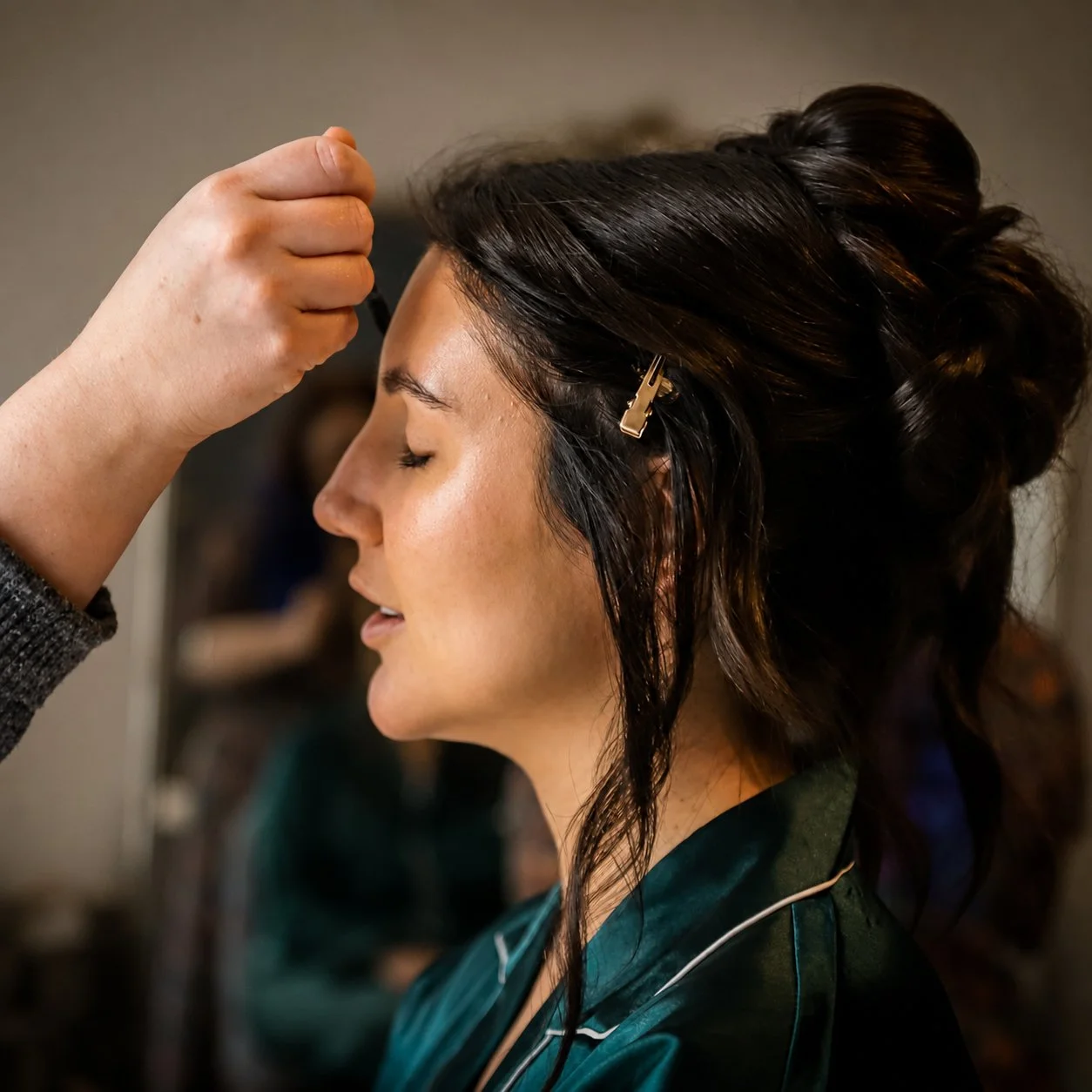 A woman with dark, wavy hair styled in an updo, wearing a dark green satin robe, has her eyes closed while a makeup artist applies makeup or styling her hair, with a blurred background.