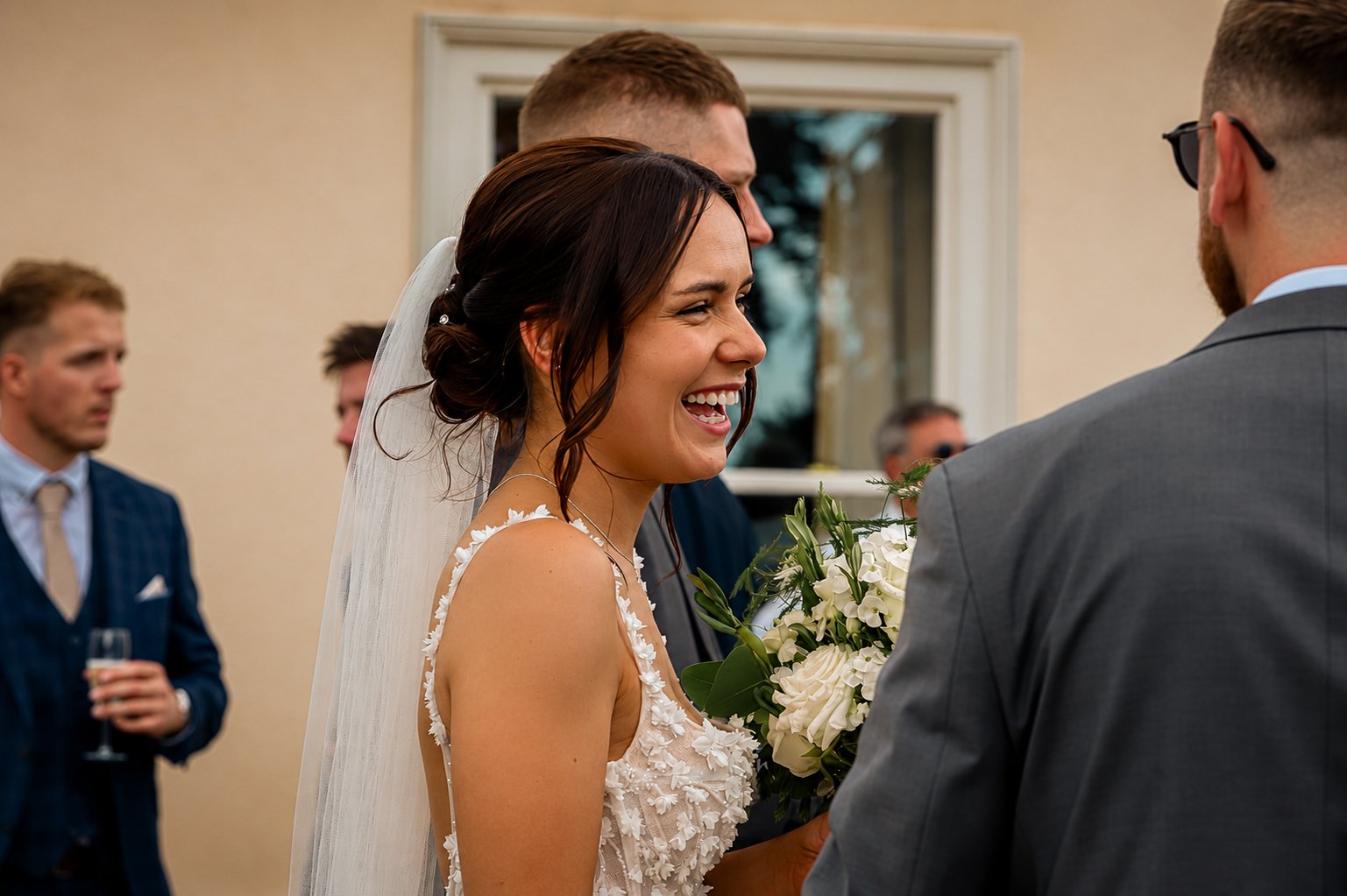 A bride with dark hair styled in loose curls, wearing a white wedding dress with floral appliqués and a veil, smiling and holding a bouquet of white flowers as she looks at a groom during a wedding ceremony. In the background, groomsmen are visible, 