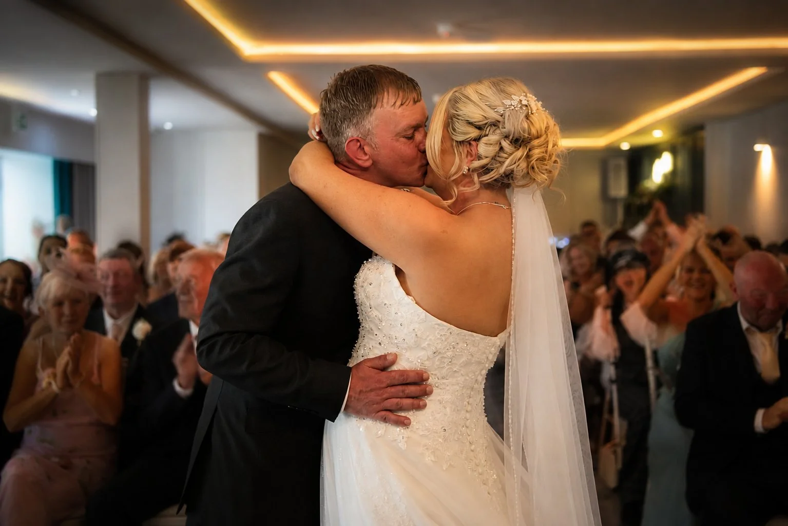 A bride and groom share a kiss during their wedding reception, surrounded by seated guests clapping and smiling.