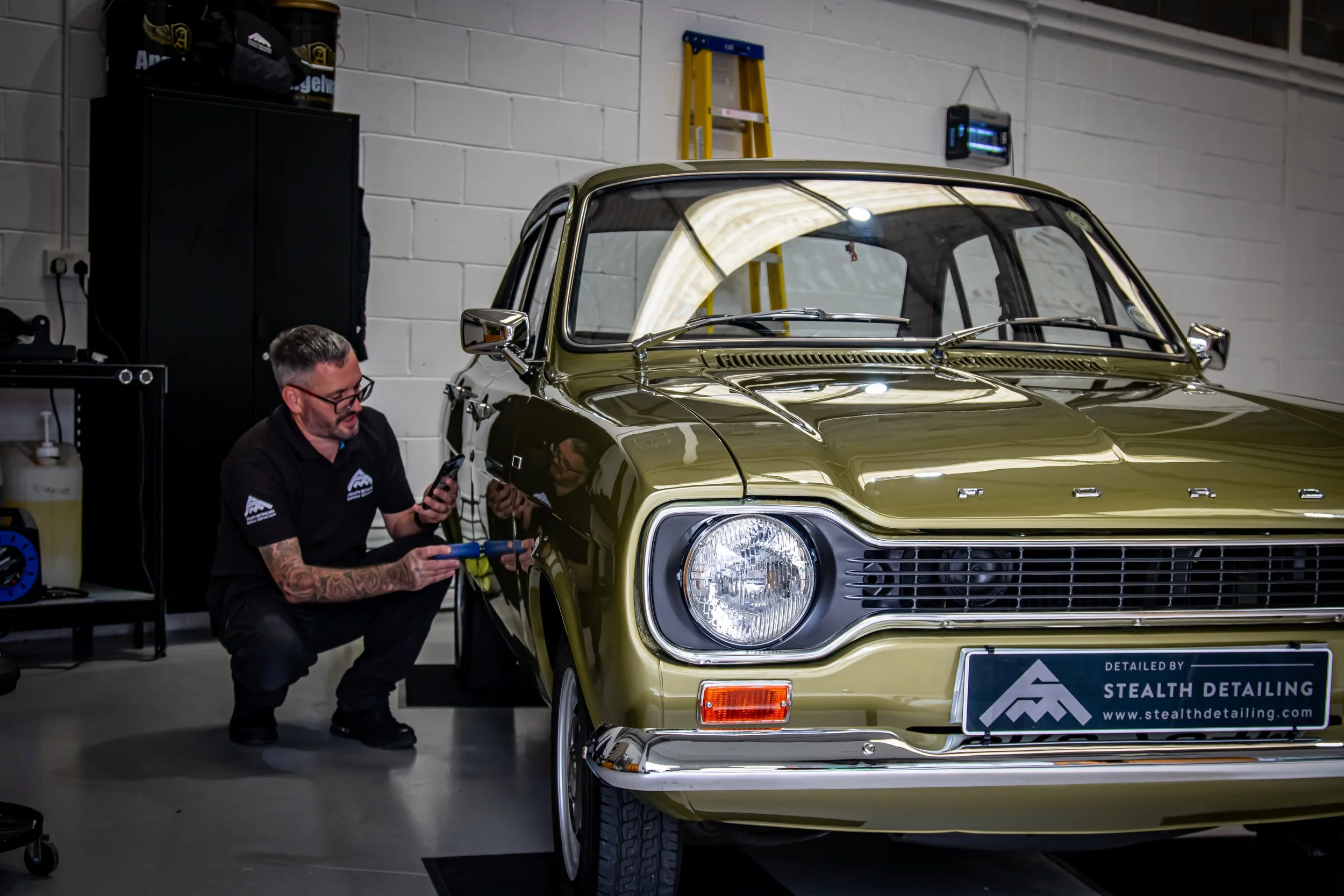 A man with tattoos and glasses crouching next to a vintage green car, working with a tool, in an indoor garage or workshop setting.