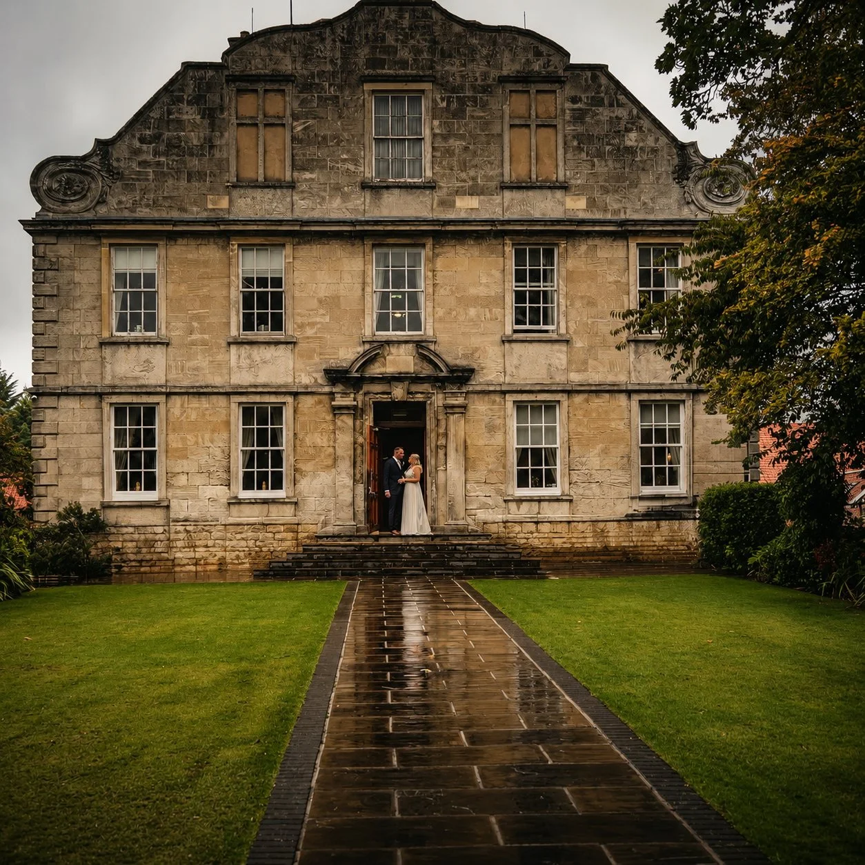 A bride and groom stand in a doorway of an old, three-story stone mansion on a rainy day, holding hands and looking at each other. The mansion has multiple windows, some of which are boarded up, and the scene is framed by green grass and trees.