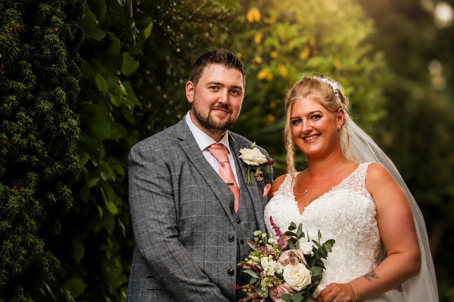 A newlywed couple standing outdoors, smiling at the camera. The groom wears a gray checked suit with a pink tie and a white rose boutonnière. The bride wears a white lace wedding dress and veil, holding a bouquet of flowers, with long blonde hair sty