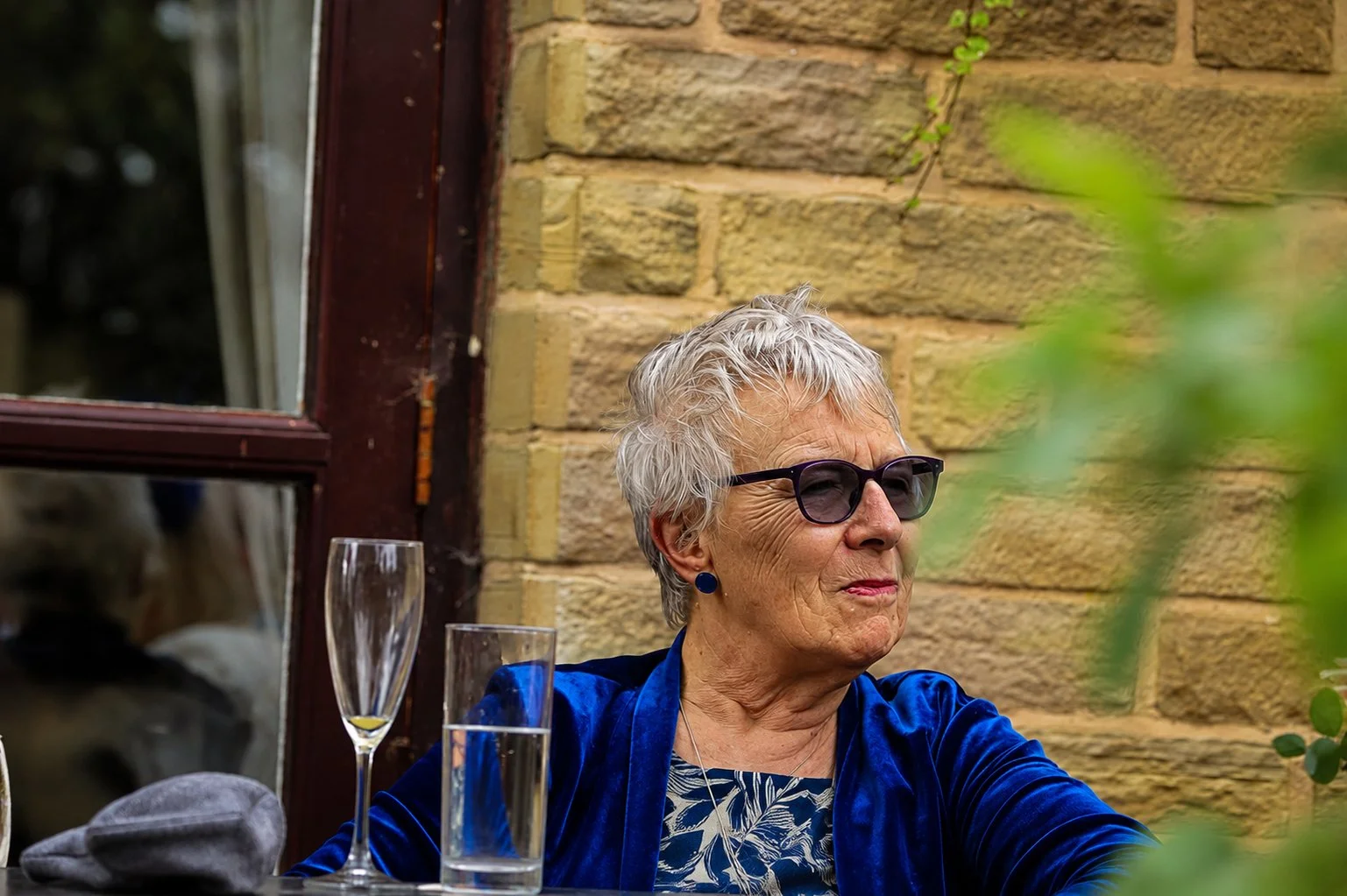 An elderly woman with short gray hair, wearing glasses, earrings, and a blue jacket, sitting outdoors at a table with an open window and a brick wall in the background. There are a wine glass, a water glass, and a gray hat on the table.