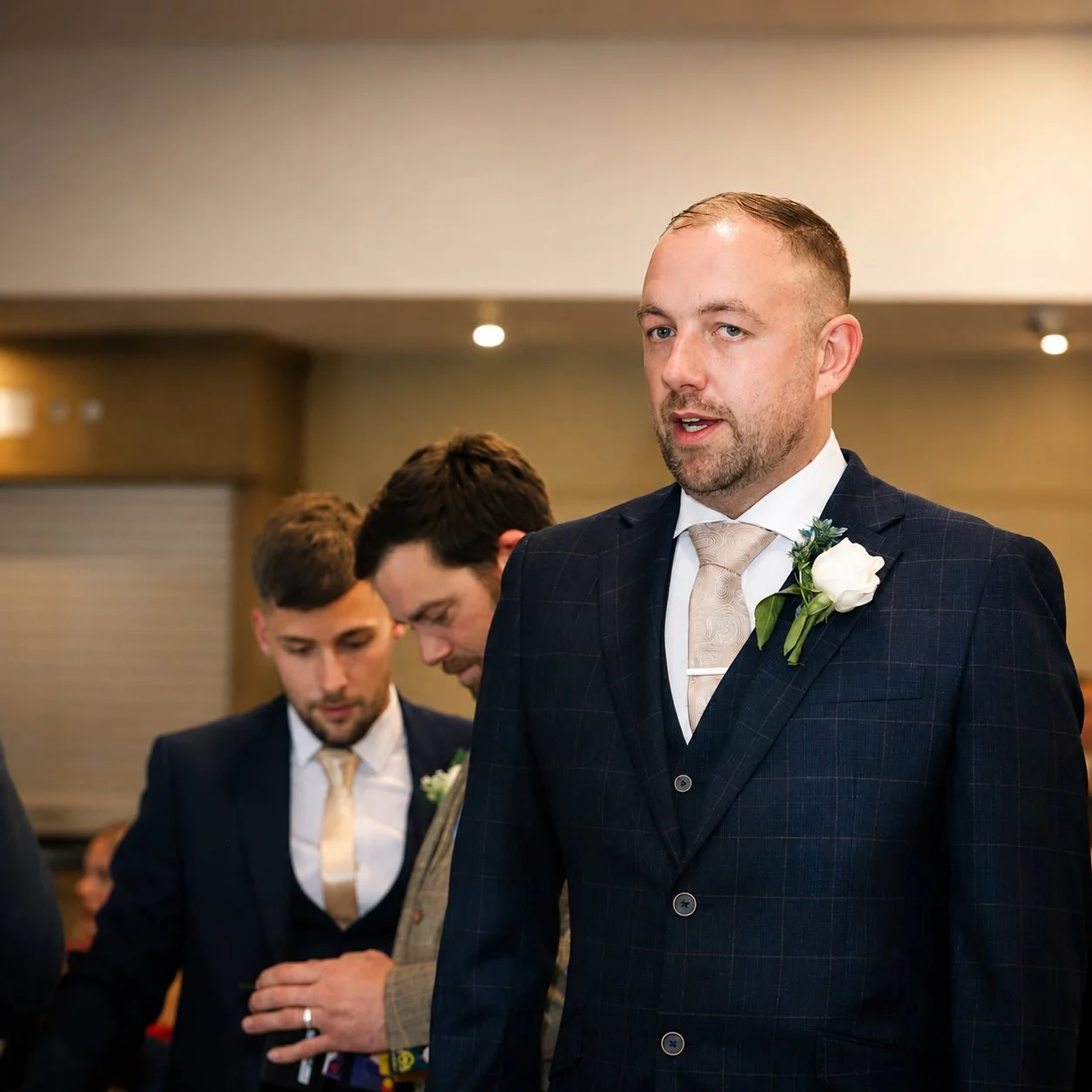 A man in a dark suit with a white boutonniere, speaking during a formal event, with two men in similar attire looking down in the background.