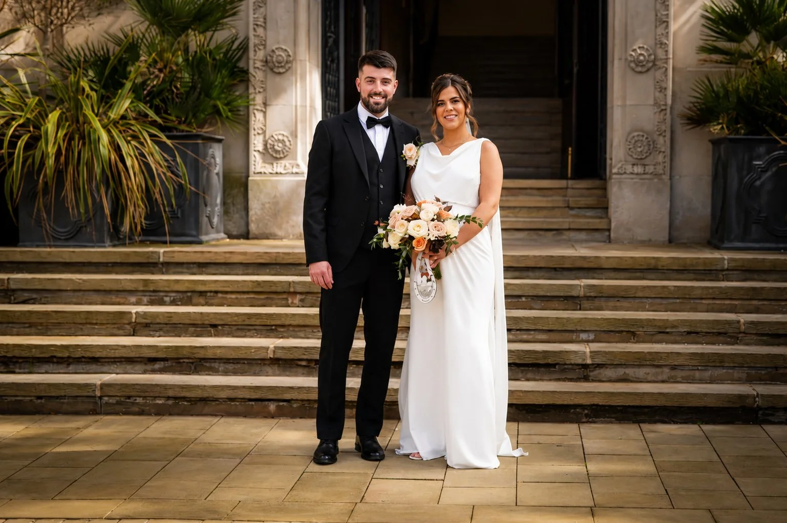 A bride and groom pose together on hospital steps outside a historic building, dressed in wedding attire, smiling and holding a bouquet of flowers.
