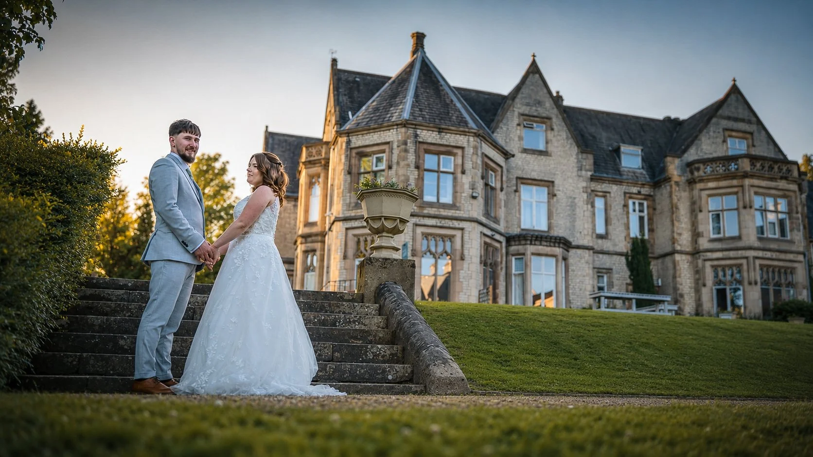 A bride and groom holding hands and smiling at each other on stone stairs outside a large stone mansion at sunset.
