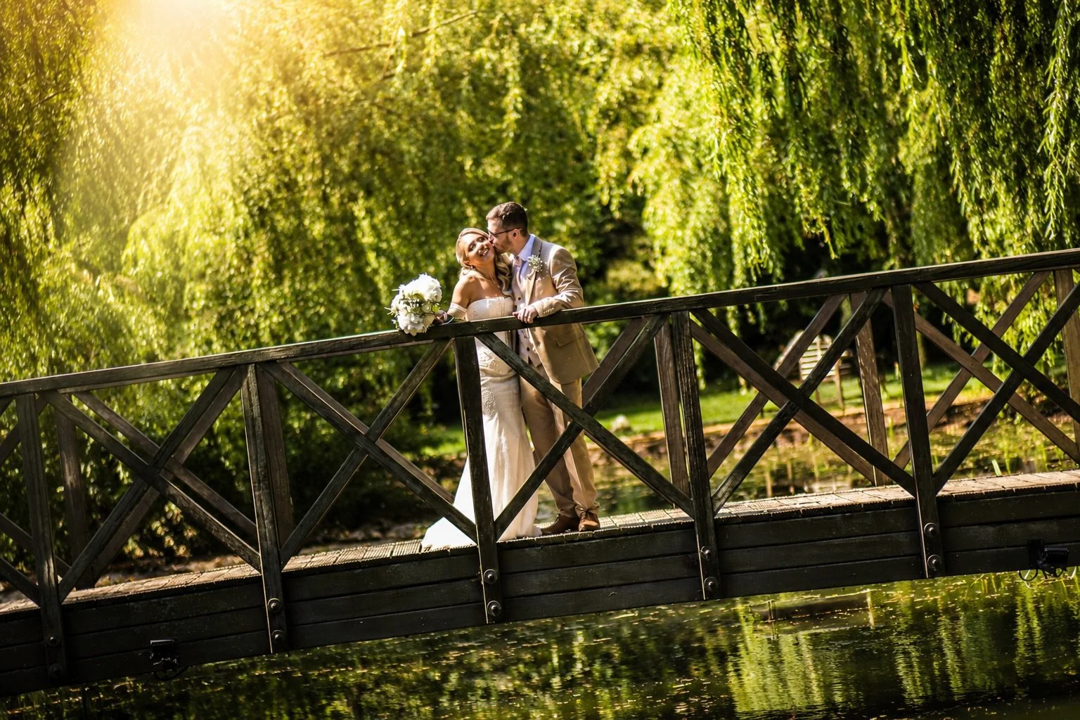 A bride and groom standing on a wooden bridge by a pond, sharing a kiss in a lush green park. The bride holds a bouquet of white flowers, and sunlight filters through the trees.