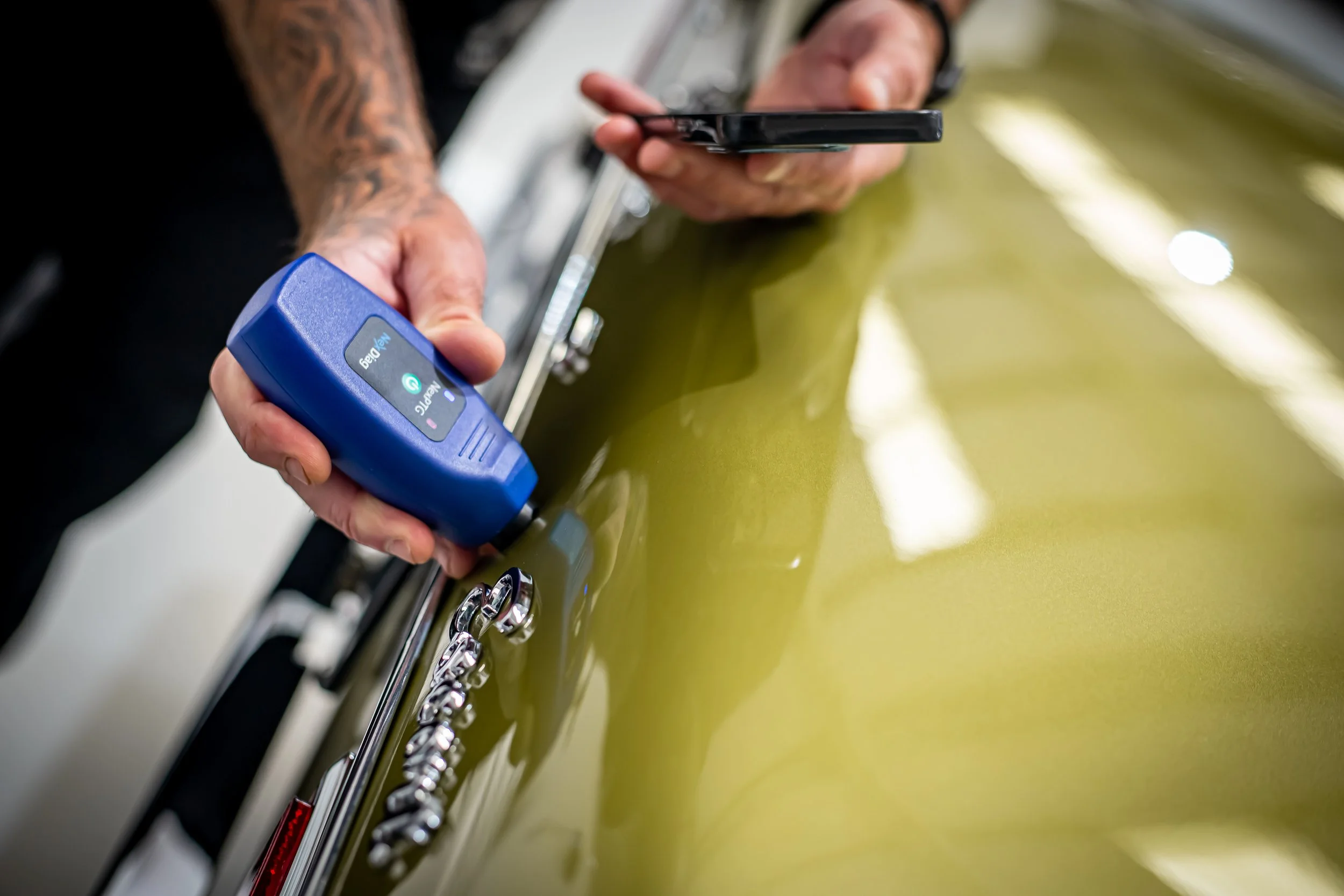 Close-up of a person in a black shirt using a metal scraper to remove residue from a shiny yellow-green car surface. The person's arms are tattooed, and they are holding a smartphone in one hand and a blue device labeled 'Vape' in the other.