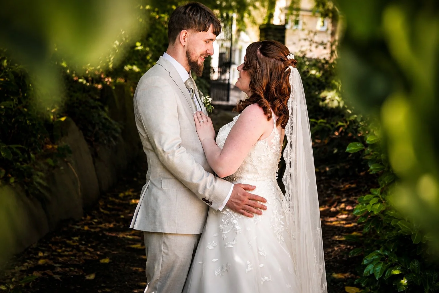A newlywed couple gazes lovingly at each other during their wedding in an outdoor garden, with the bride wearing a lace wedding dress and veil and the groom in a light-colored suit.
