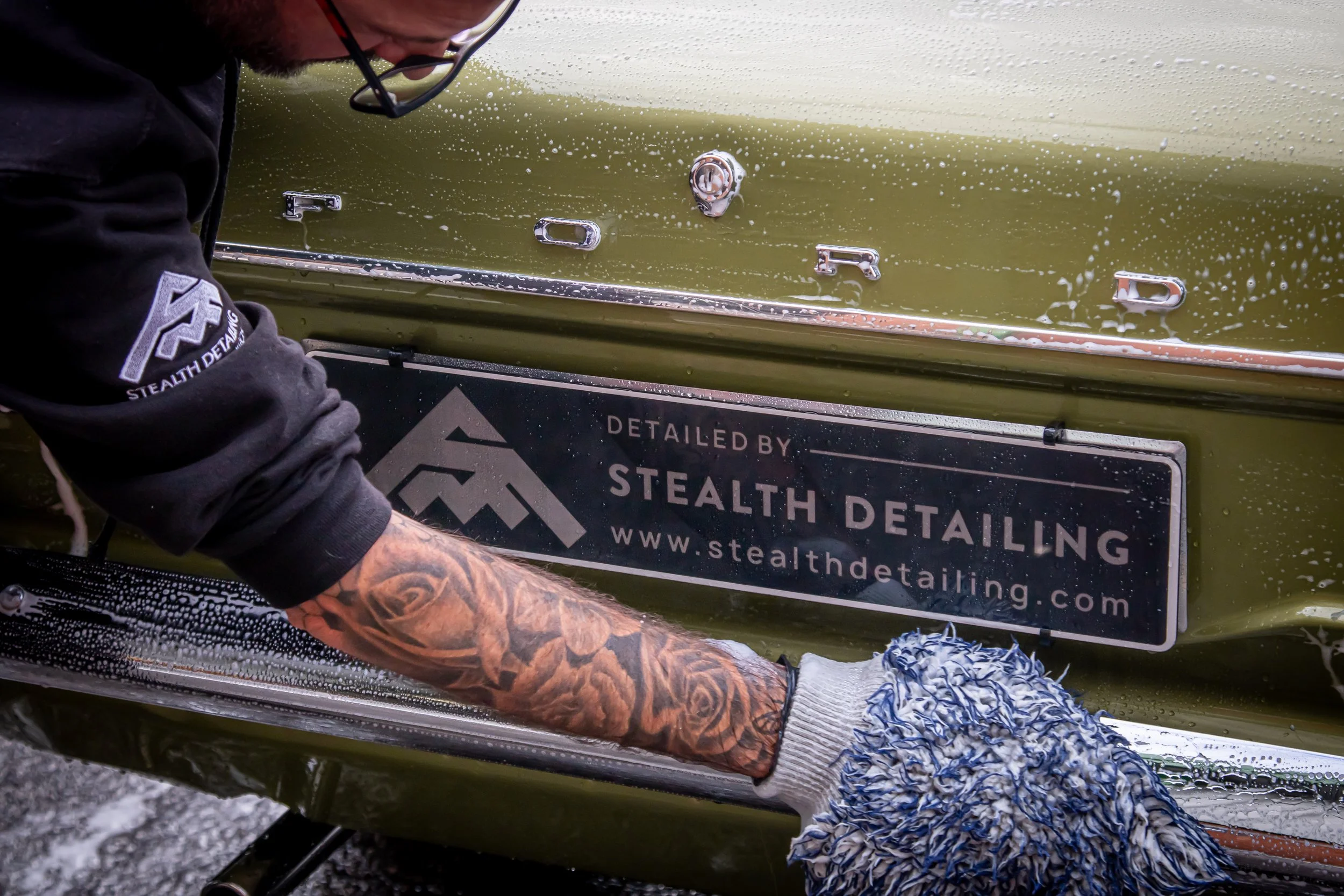 A person cleaning a green car with a sponge, water, and soap. Focus on the person's arm with tattoos, the car's faded yellow-green paint, and the black sign with white text reading 'STEALTH DETAILING.'